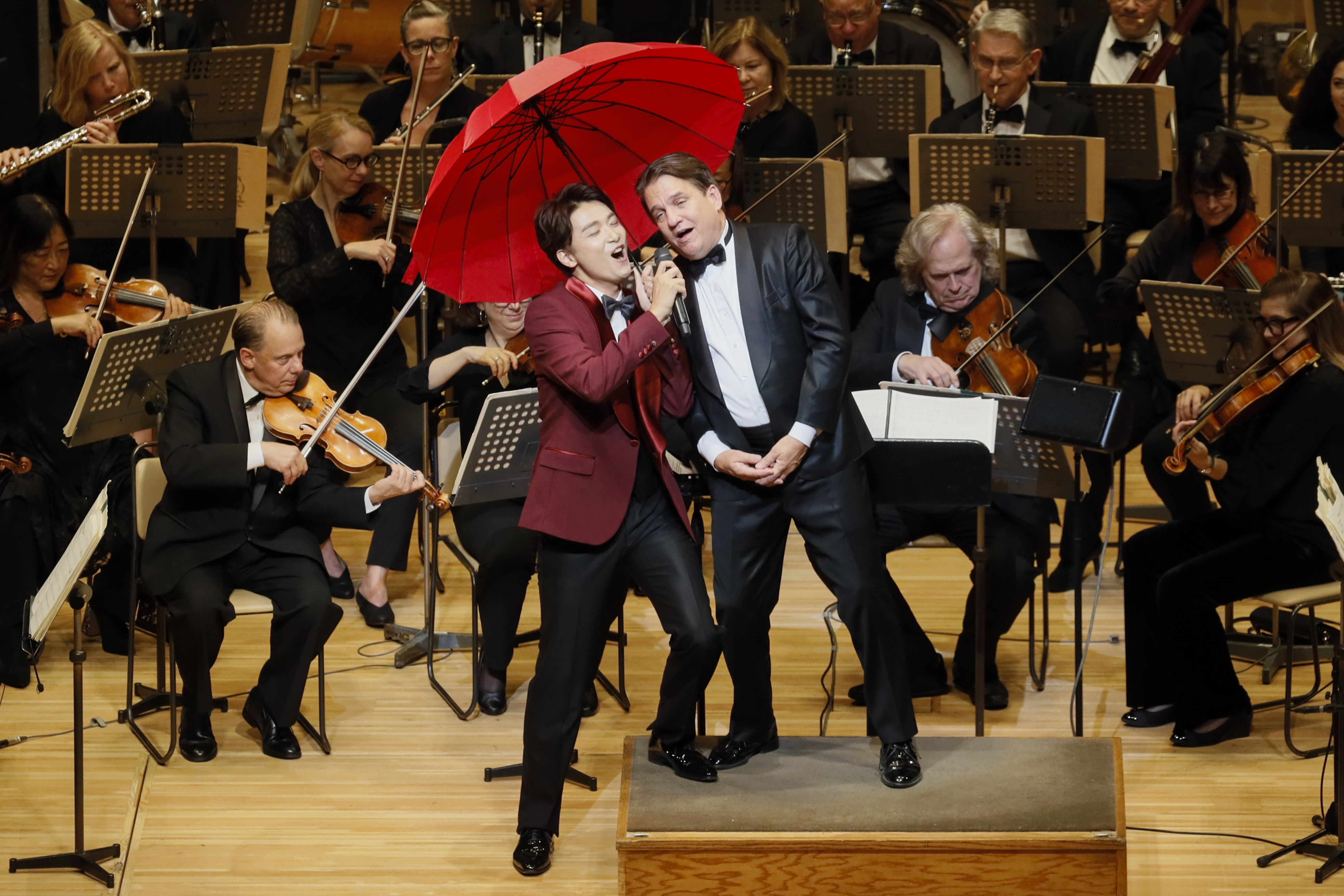 Singer Yoshio Inoue and Keith Lockhart lean together beneath a red umbrella on stage in Tokyo, Japan