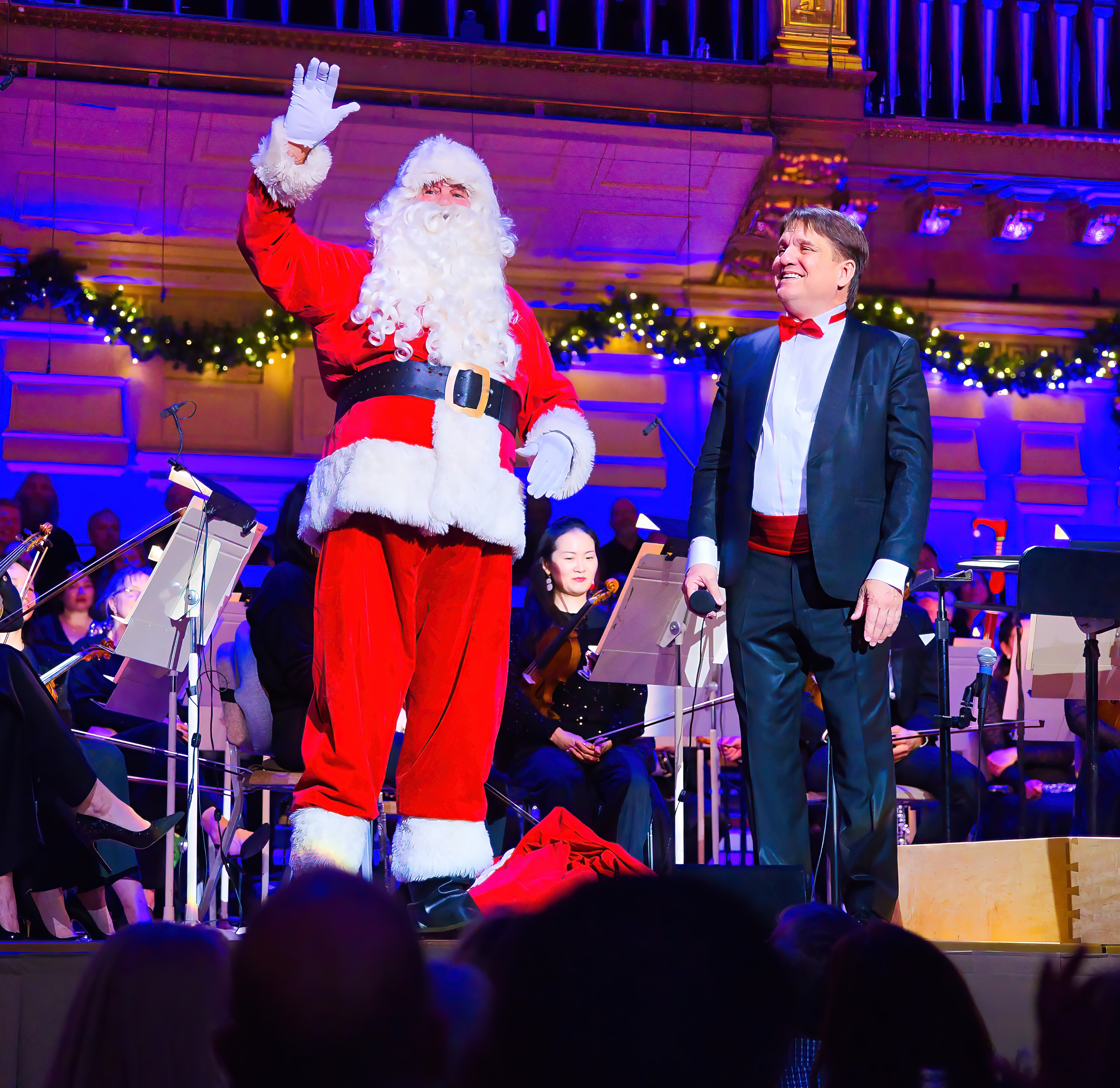 Keith Lockhart standing on the Symphony Hall stage looking at Santa Clause who is waiving at the audience