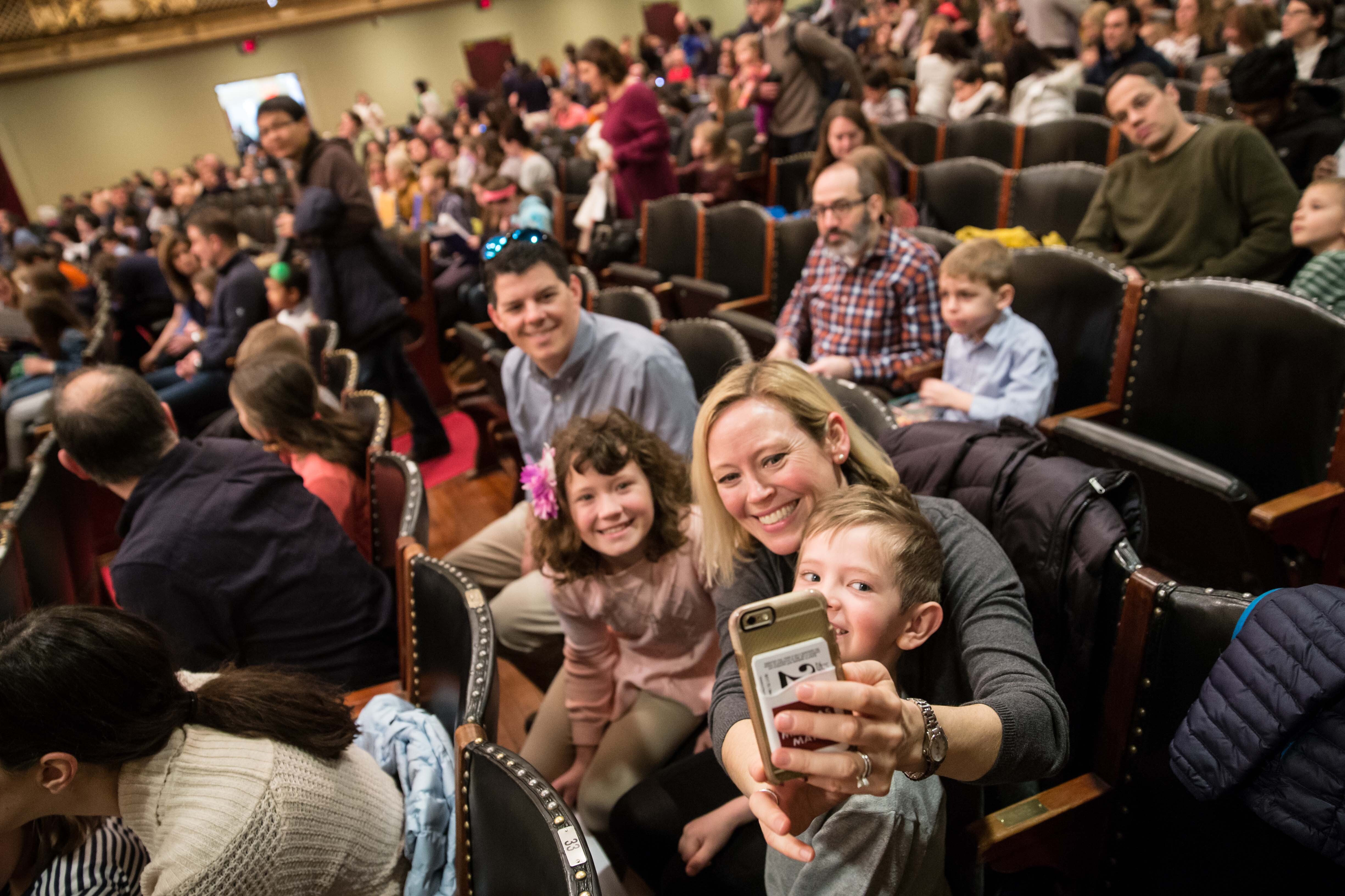 A woman with blonde hair takes a selfie with a man and two children as they sit in Symphony Hall
