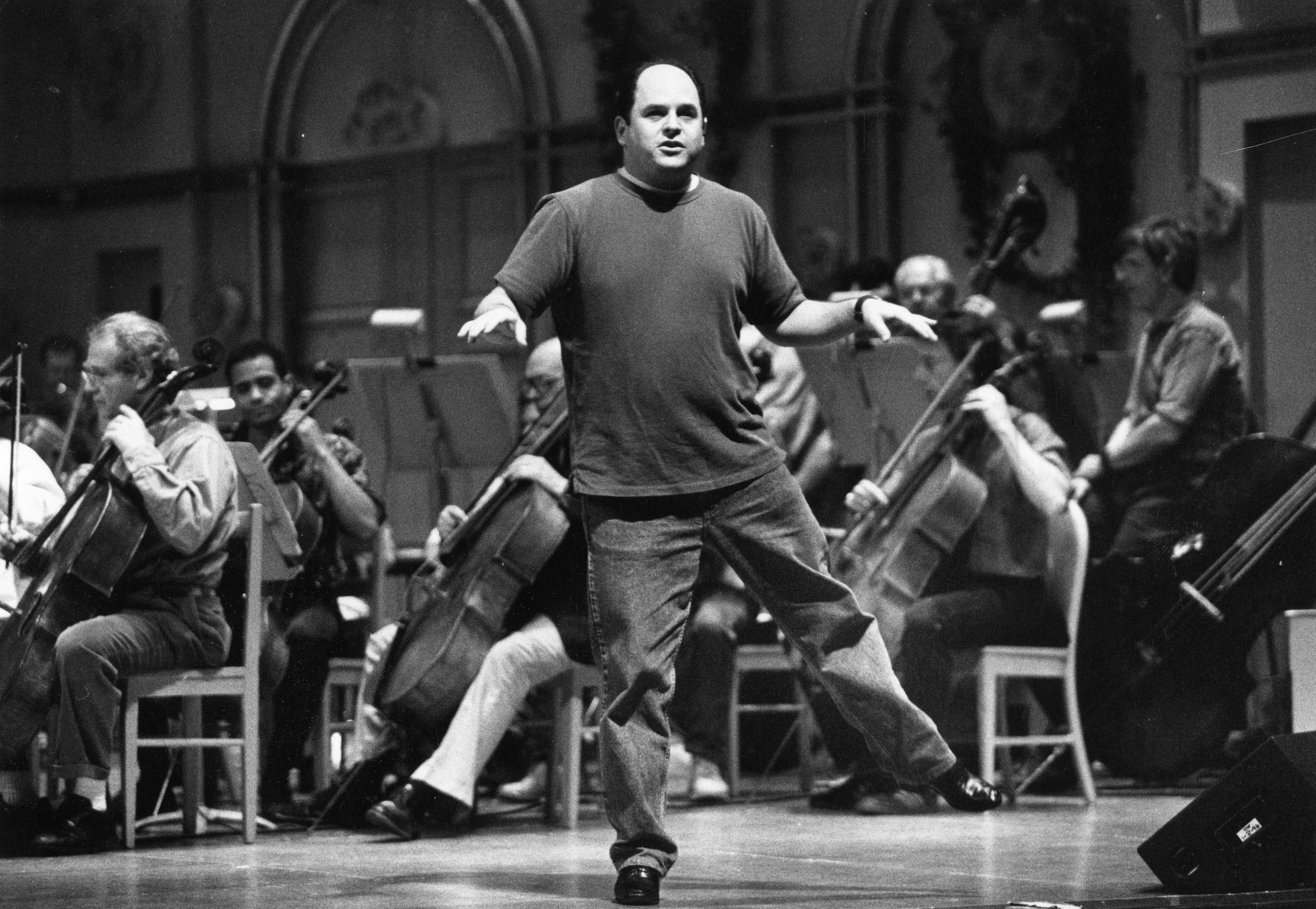 Wearing casual pants and t-shirt, guest soloist Jason Alexander poses on one leg while the orchestra plays in the background