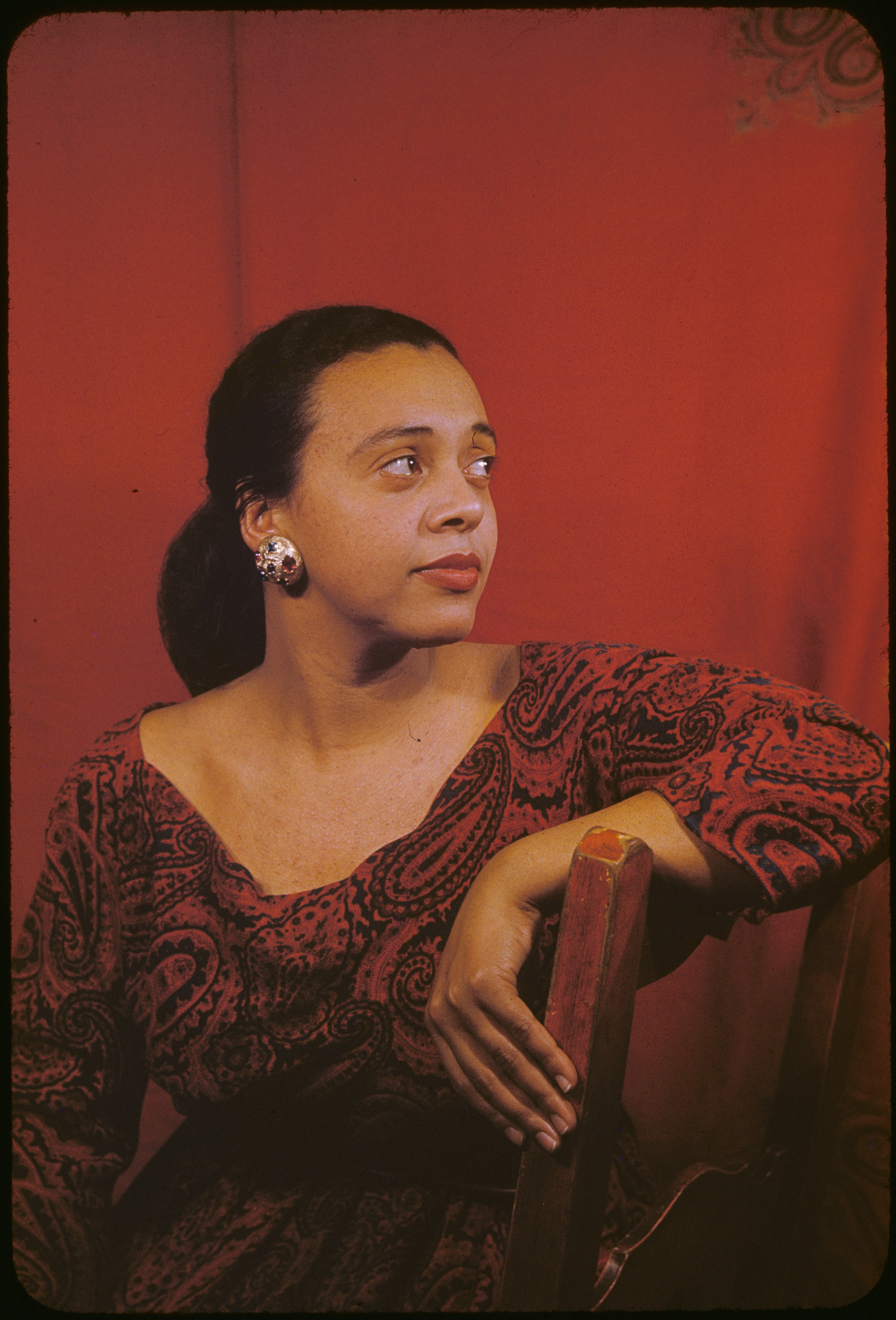 Black soprano Adele Addison is seated with her arm draped over the chair while she gazes off screen towards the right. She is wearing a red and black dress with a red backdrop behind her.