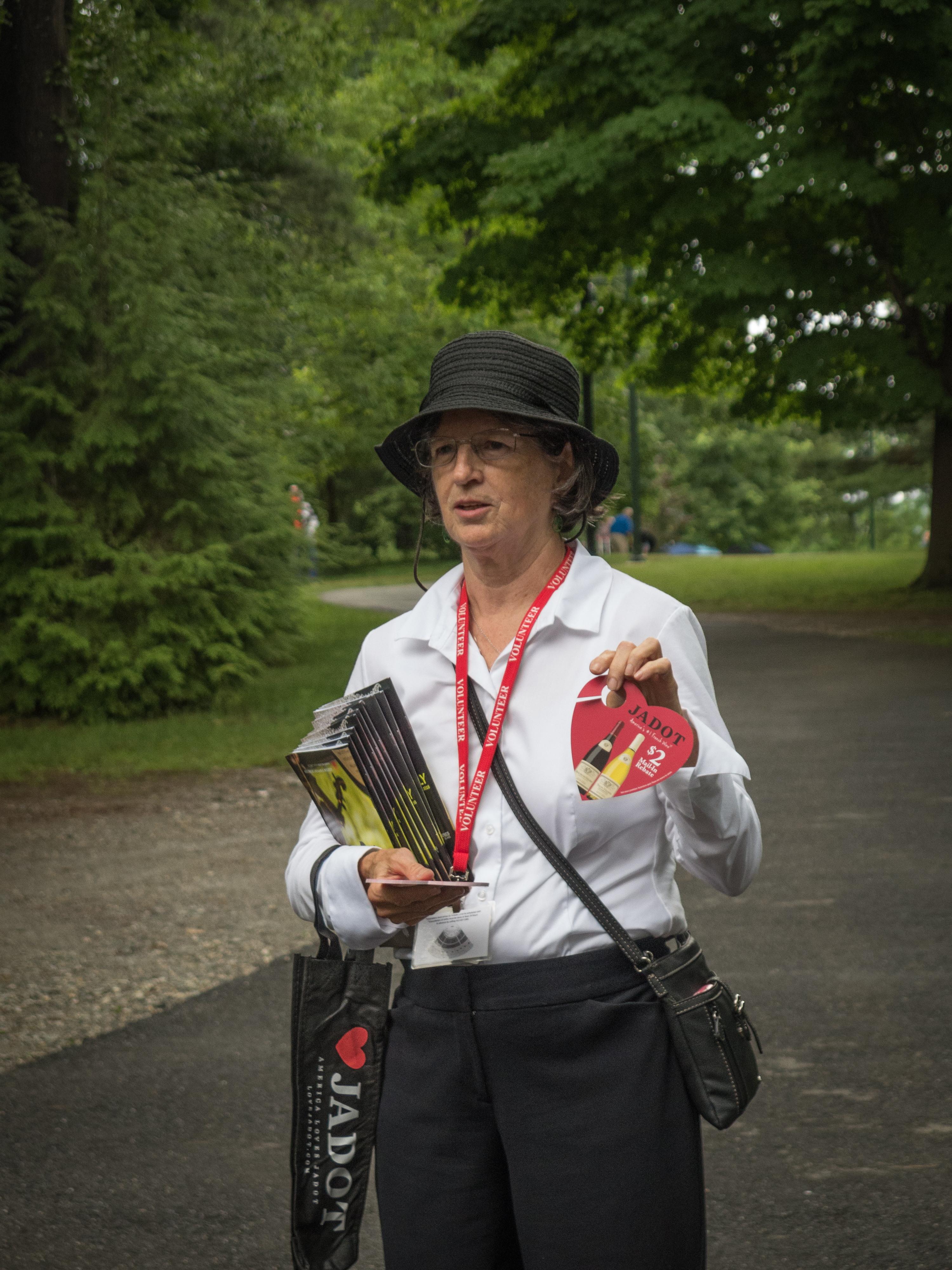 Woman wearing black hat and white shirt with red lanyard holds a stack of programs while standing on outside pathway in front of trees