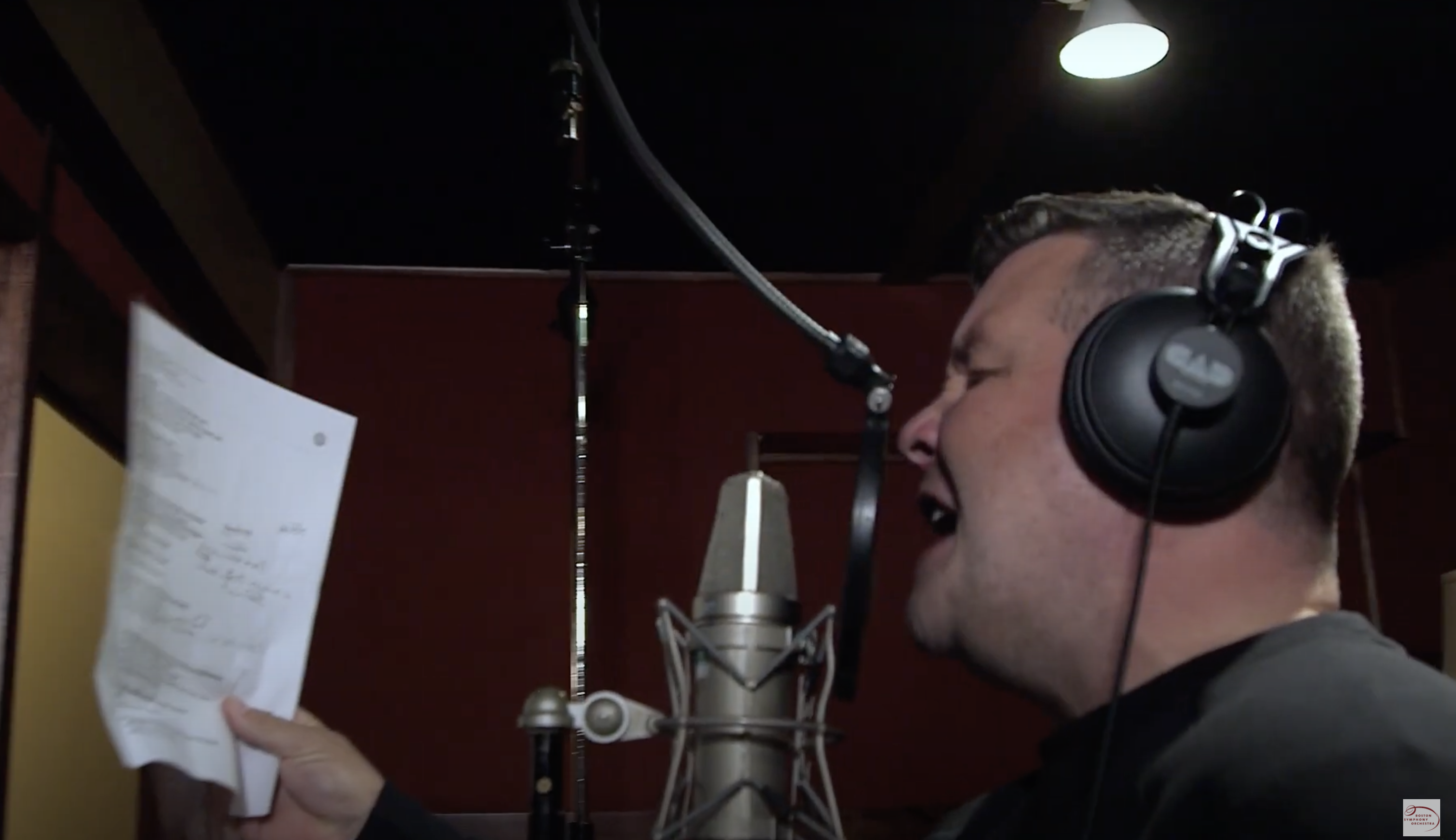 A man with a sheet of lyrics sings in a recording studio