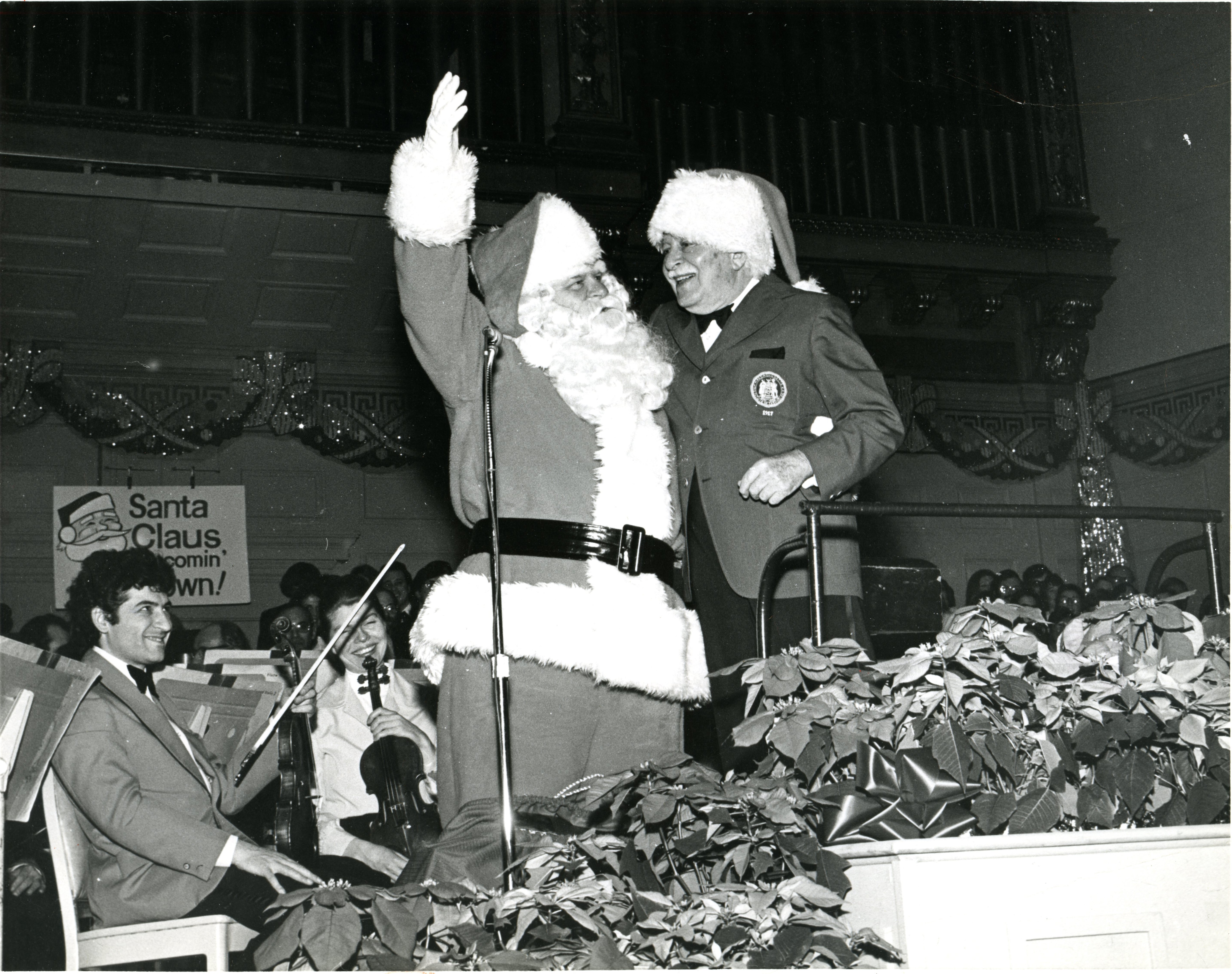 Black and white photo of a conductor and Santa Claus onstage at Symphony Hall with the Boston Pops.