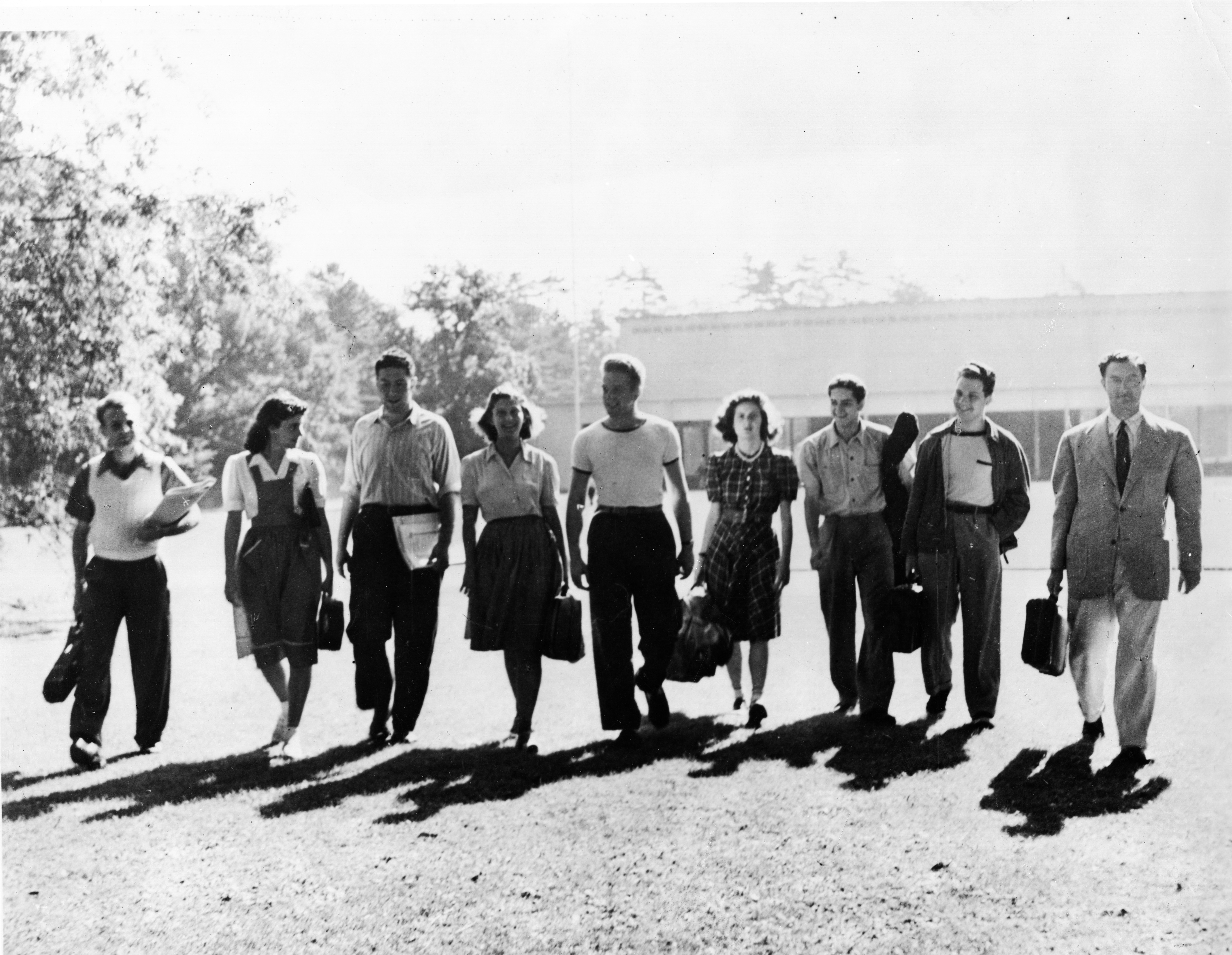 Several students holding instrument cases and wearing vintage attire walk across the Tanglewood lawn with the Music Shed in the background.