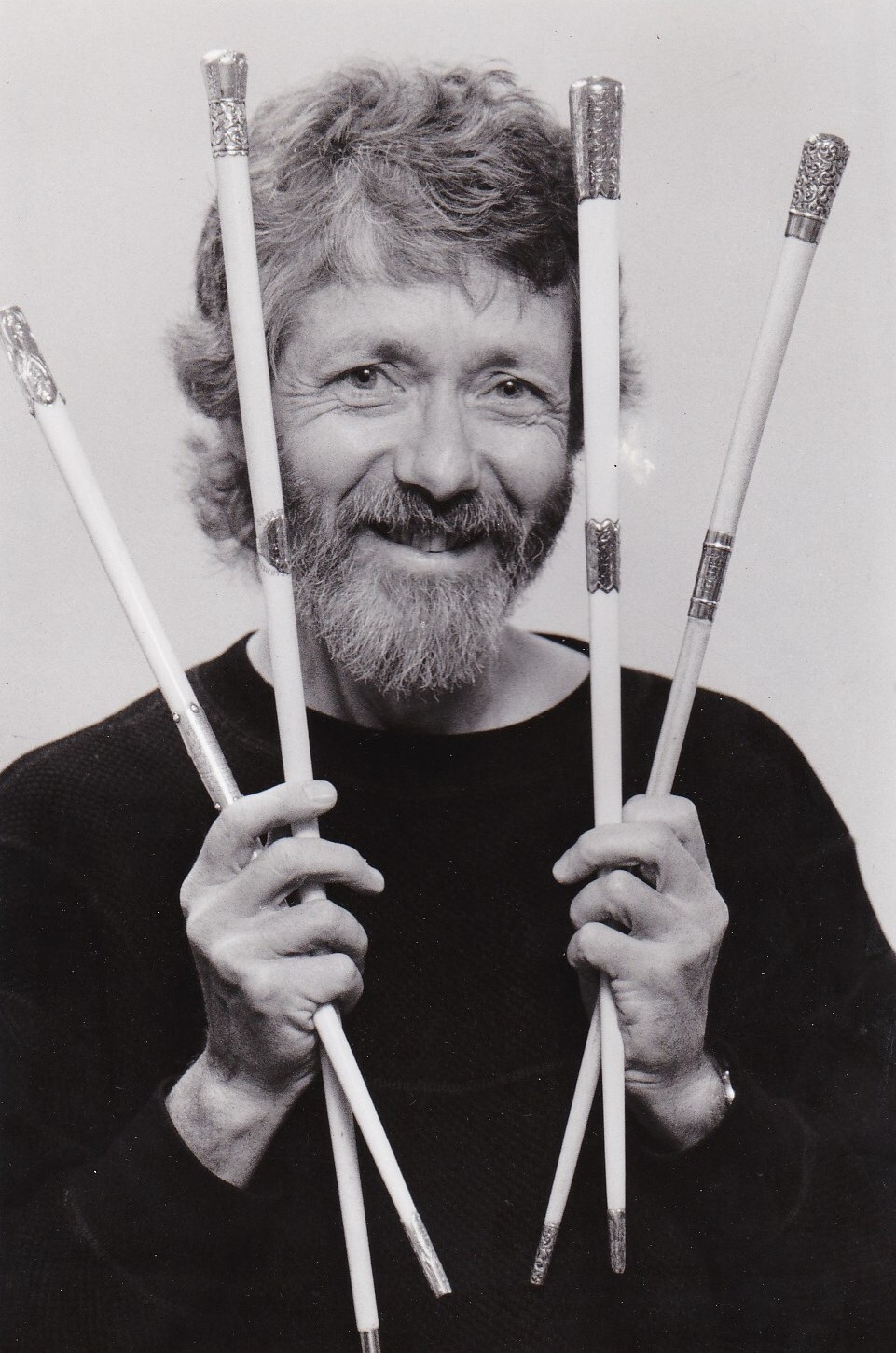 Black and white photograph of a man with beard and mustache holding up four long ivory batons with metal tips