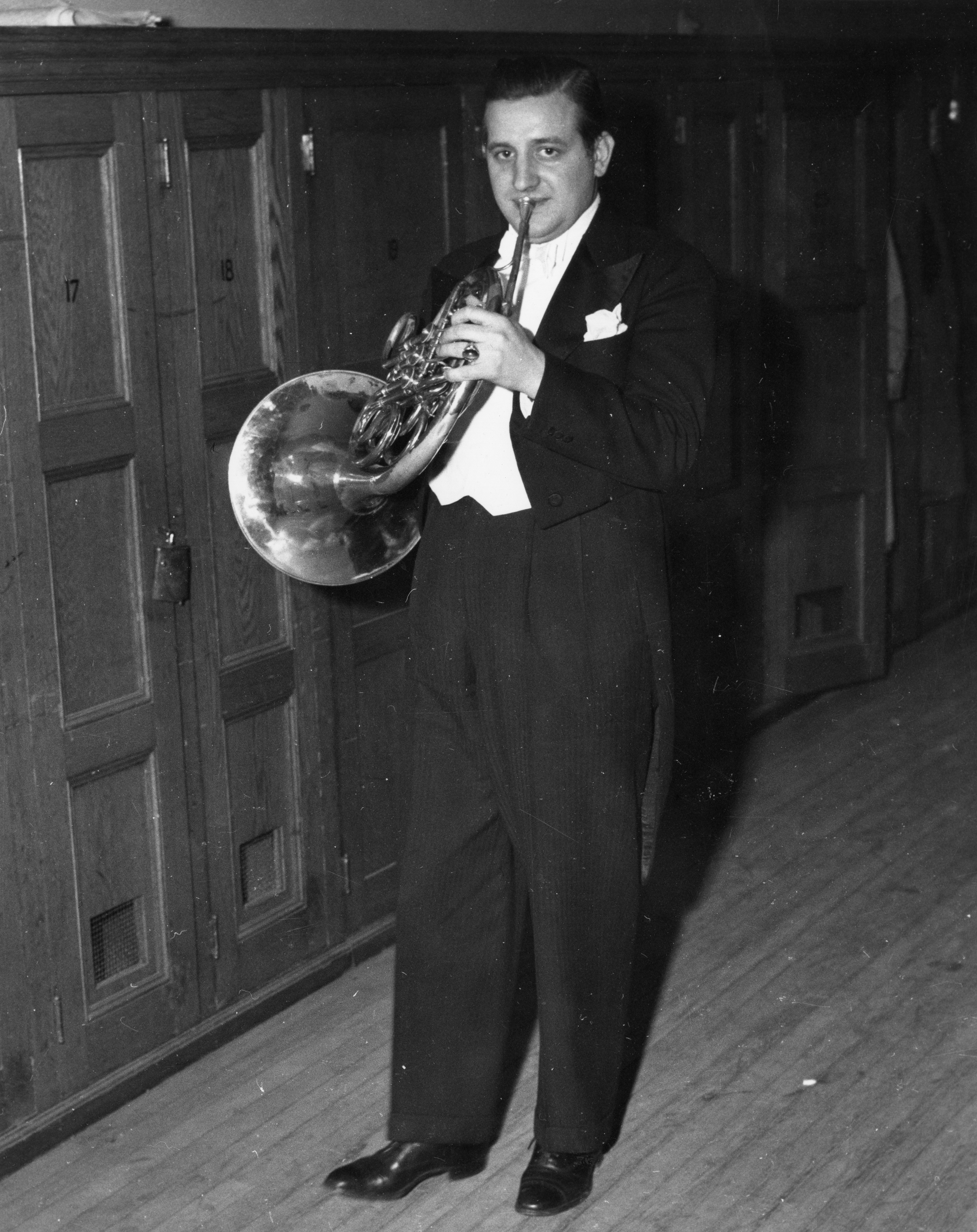 A man dressed in a tuxedo stands backstage by some wooden lockers holding his horn