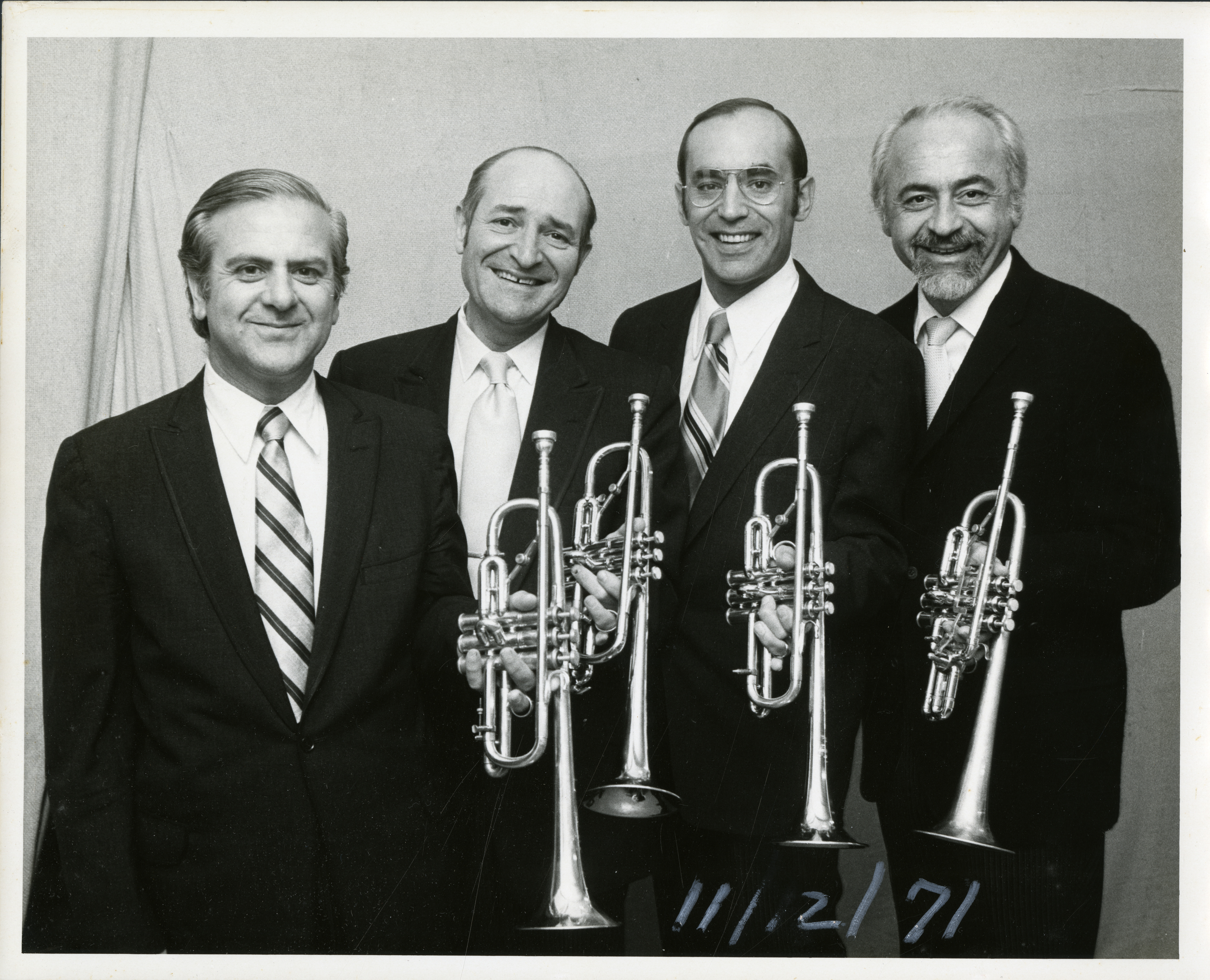 Four men stand together in suits and wide ties, each holding up a trumpet