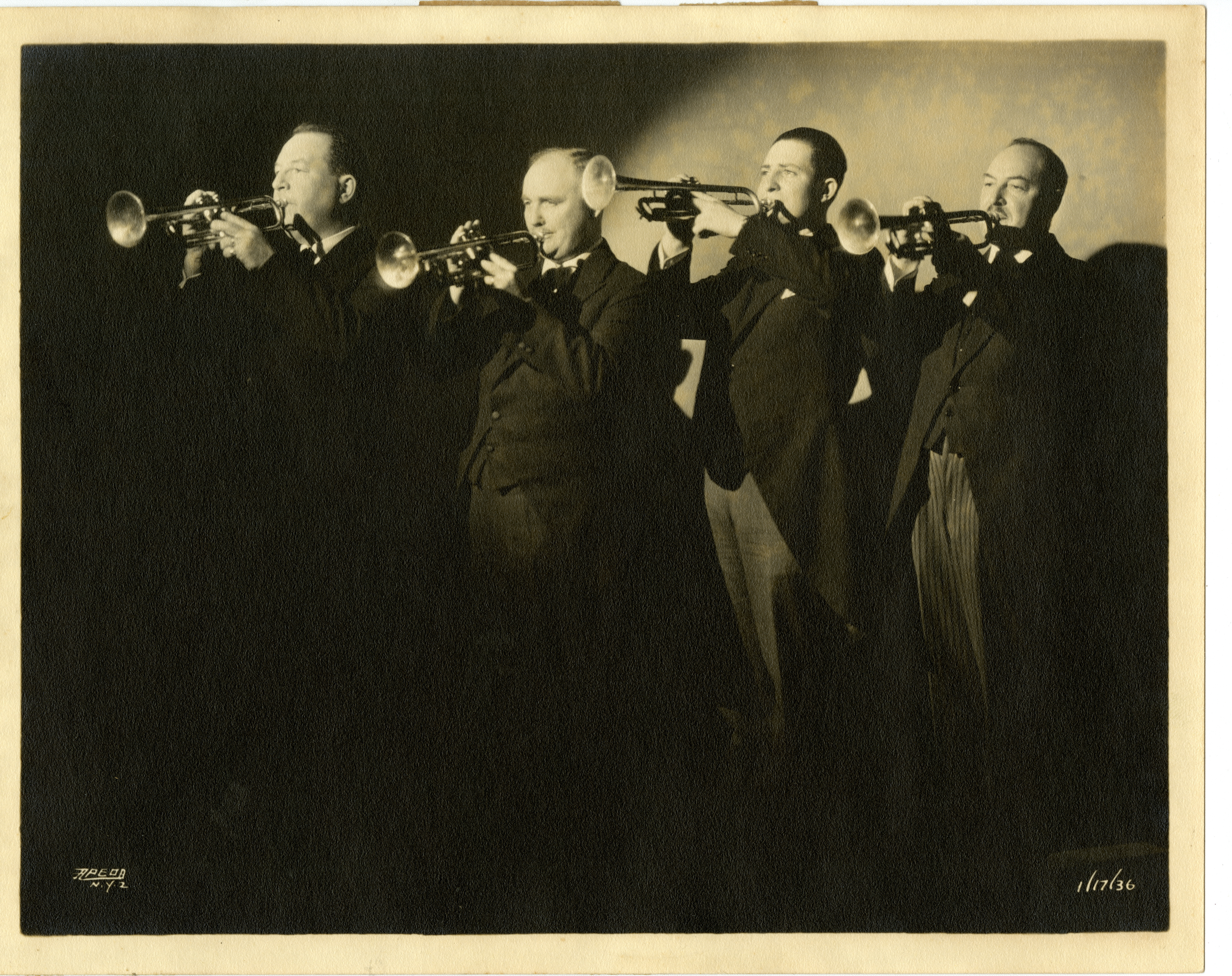 Four men in suits standing under spotlight holding their trumpets to their mouths, ready to play
