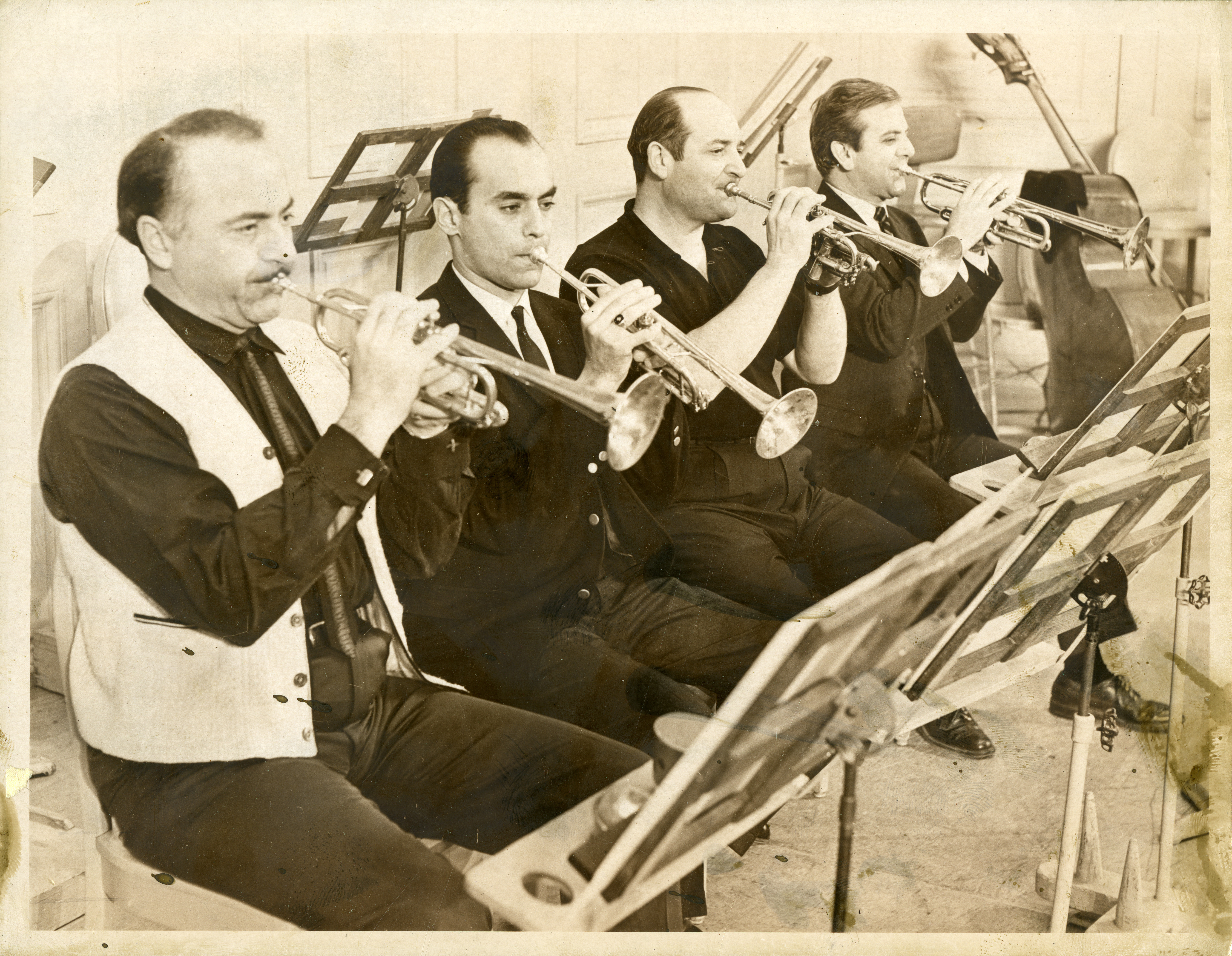 Four men in casual clothing sit in front of music stands, holding their trumpets up