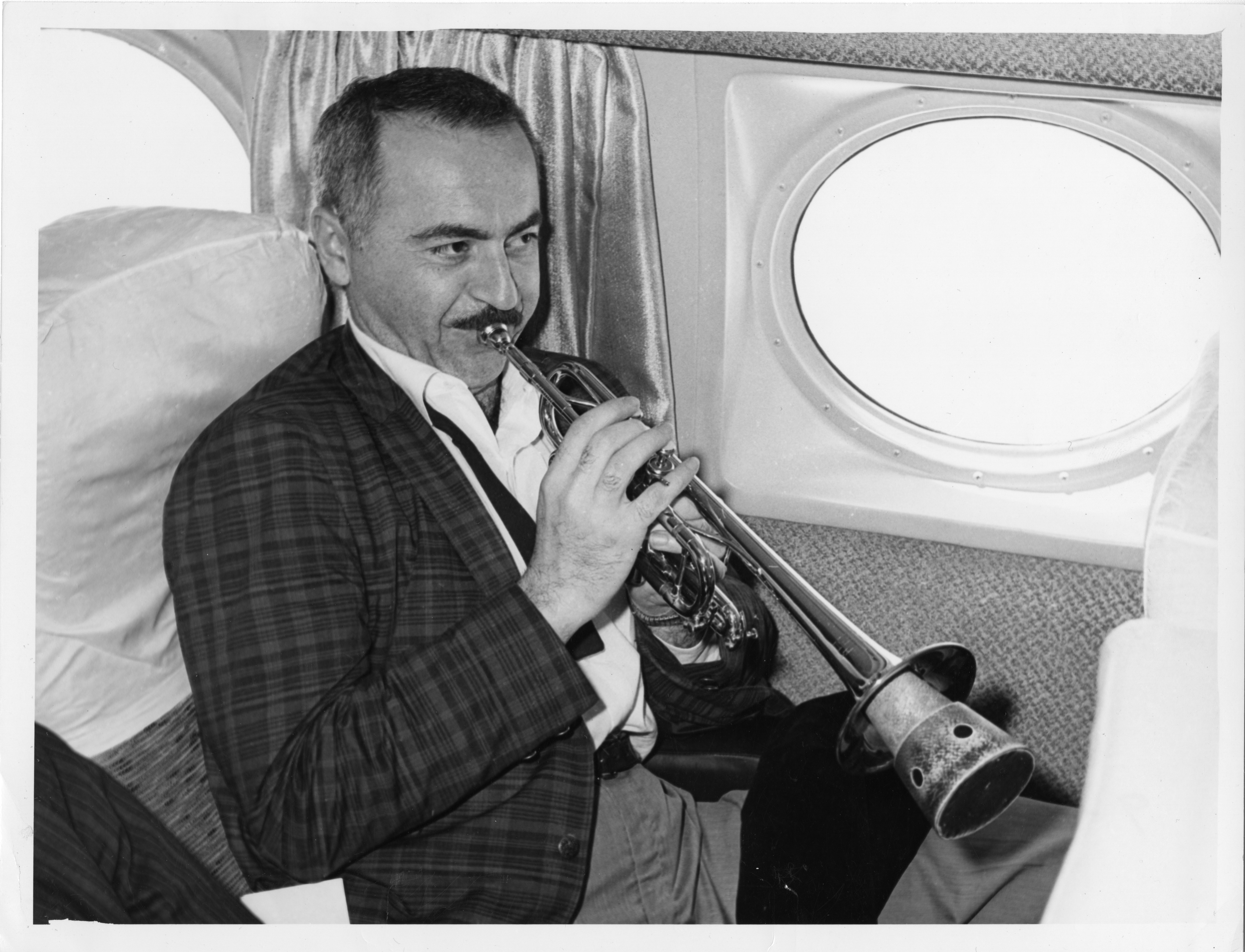 A man sits near the window of an airplane holding a trumpet with a mute fitted inside. He is wearing a suit, but the tie is unknotted, and the top buttons of his shirt are undone for a relaxed fit
