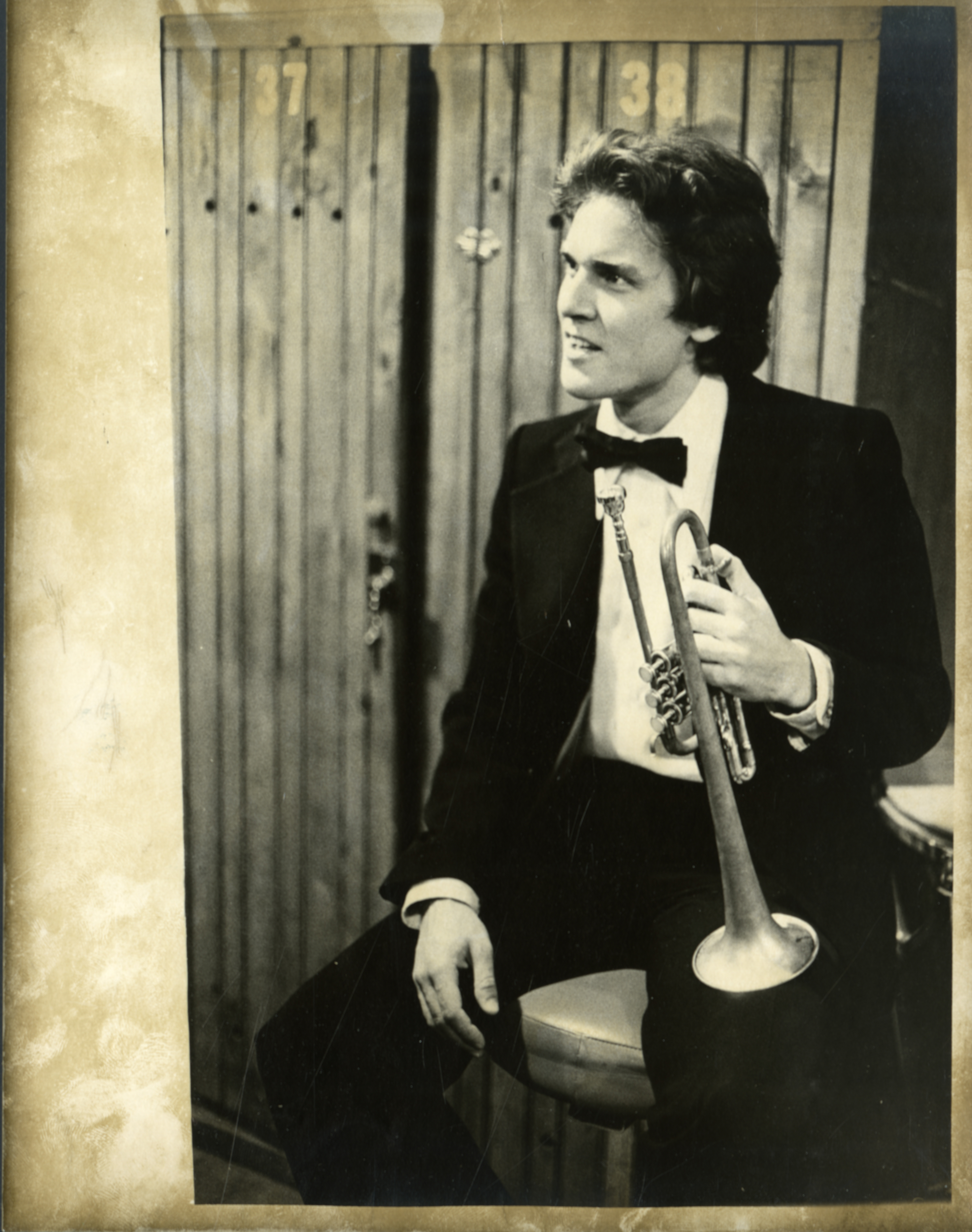 A man sits in a tuxedo and bowtie, resting a trumpet on his knee