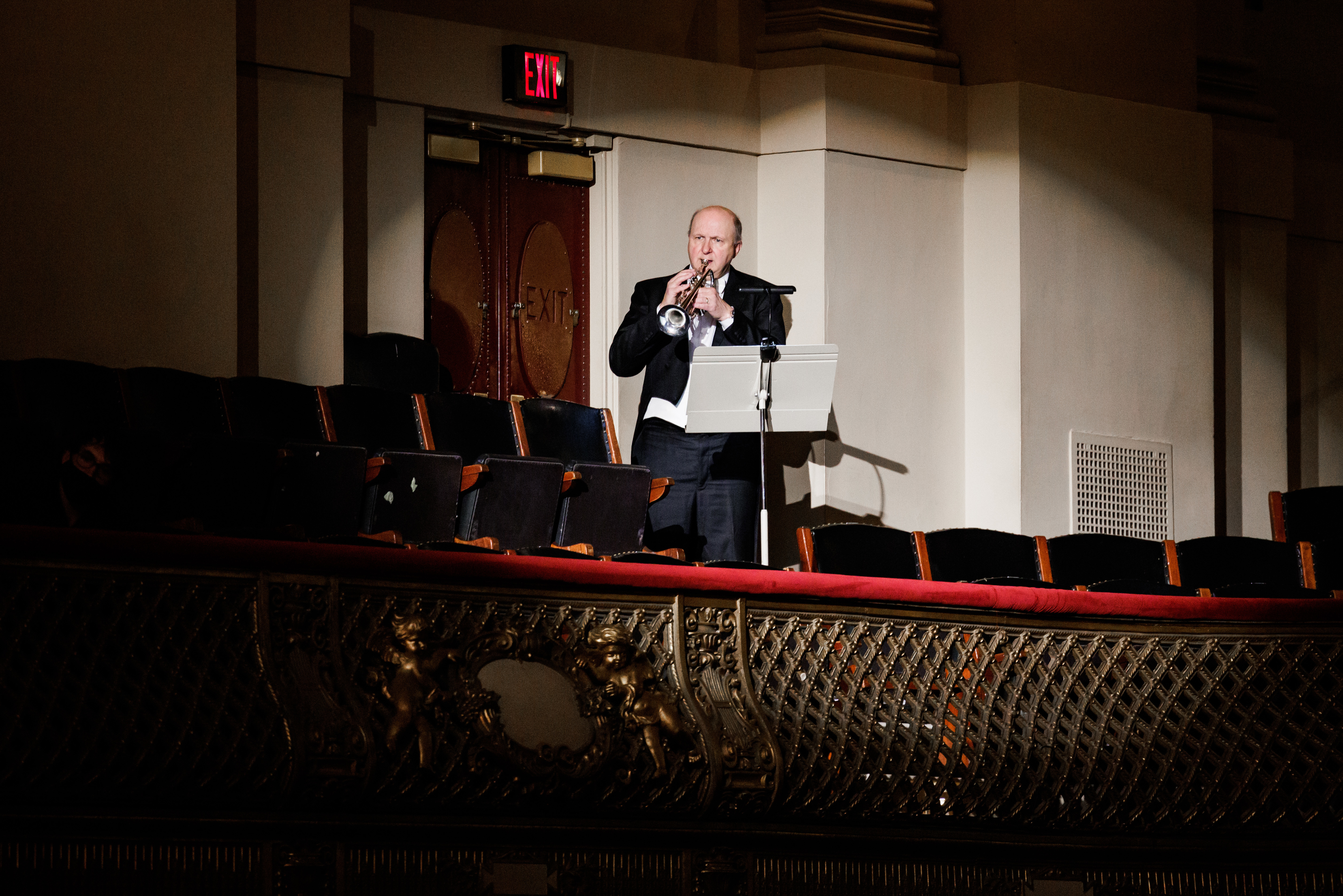 A man stands in a darkened balcony at Symphony Hall, illuminated by a spotlight while he plays a trumpet