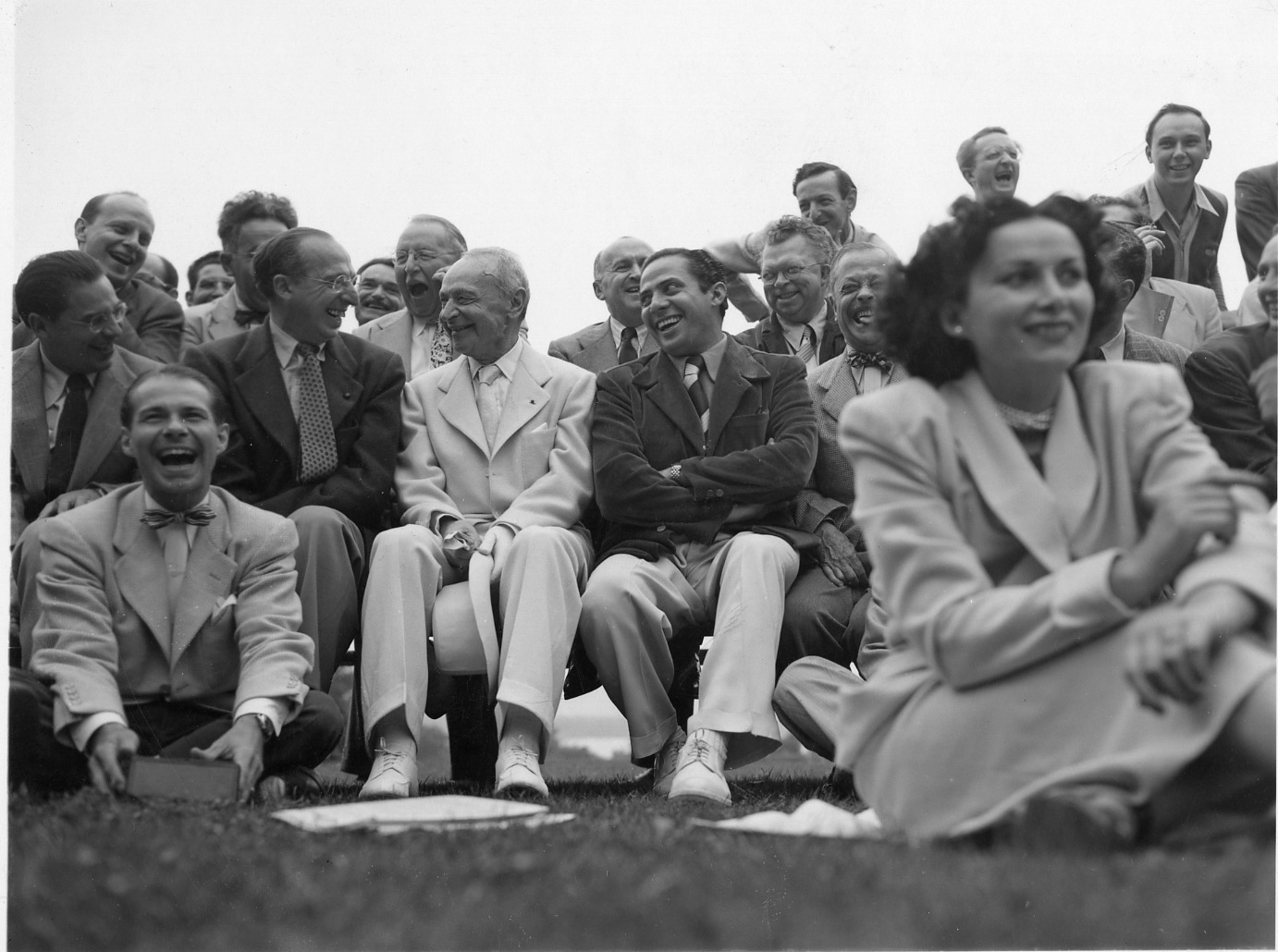 A group of laughing and smiling people (mostly men) in suits siting on benches and on the grass at Tanglewood