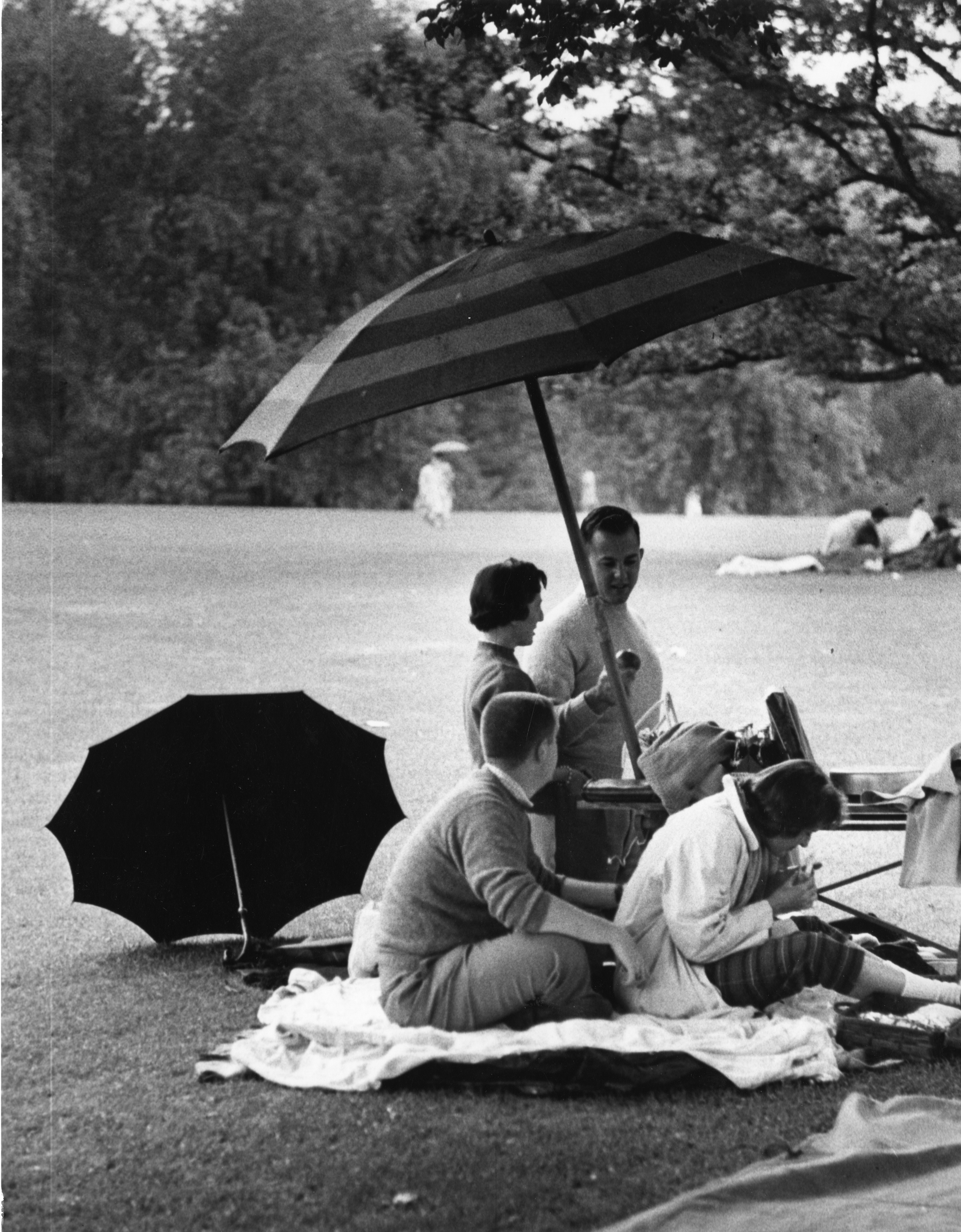 A black and white photo of four young adults sitting on a blanket, under an umbrella on the Tanglewood lawn