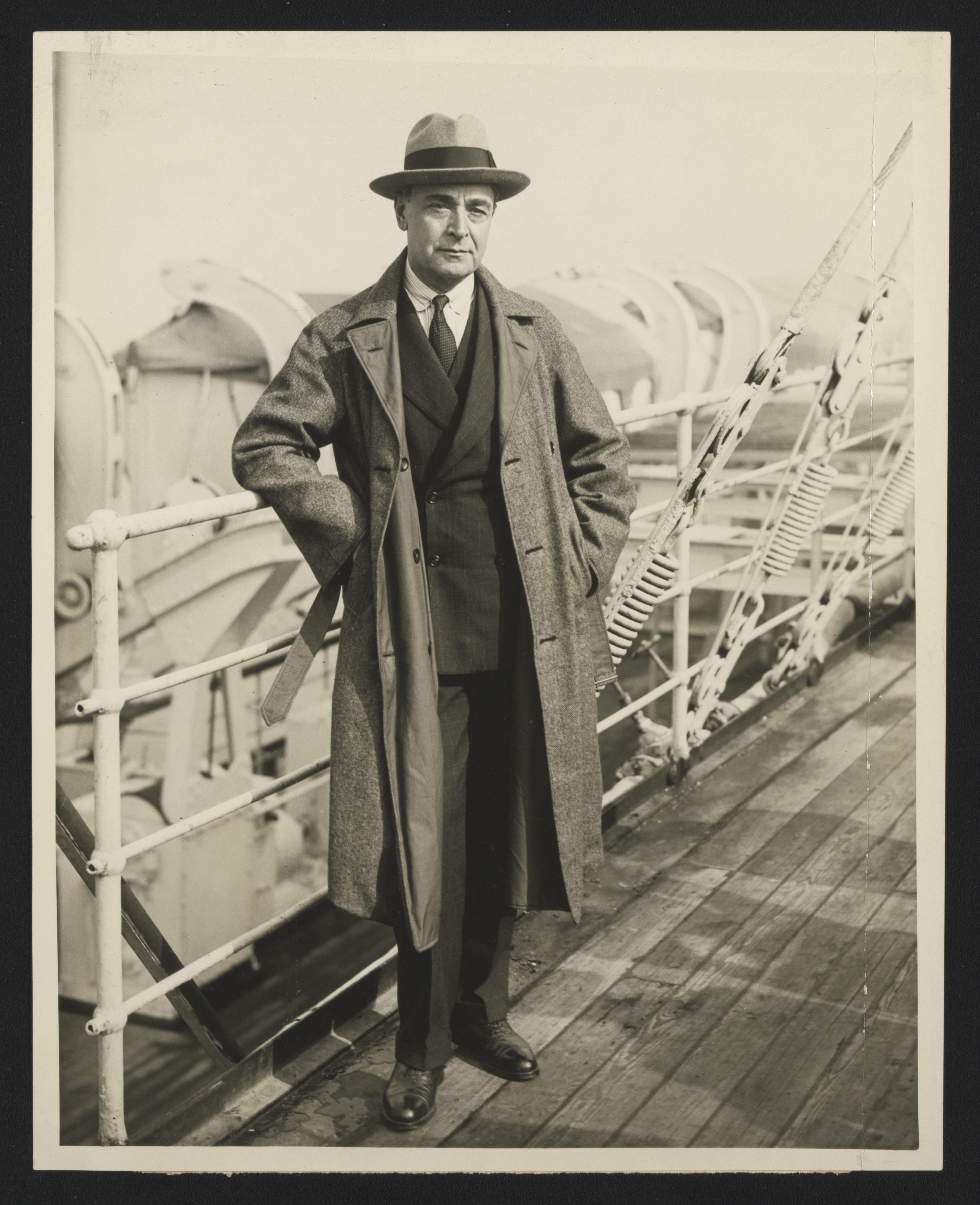 Serge Koussevitzky stands in suit, coat, and hat near a ship railing