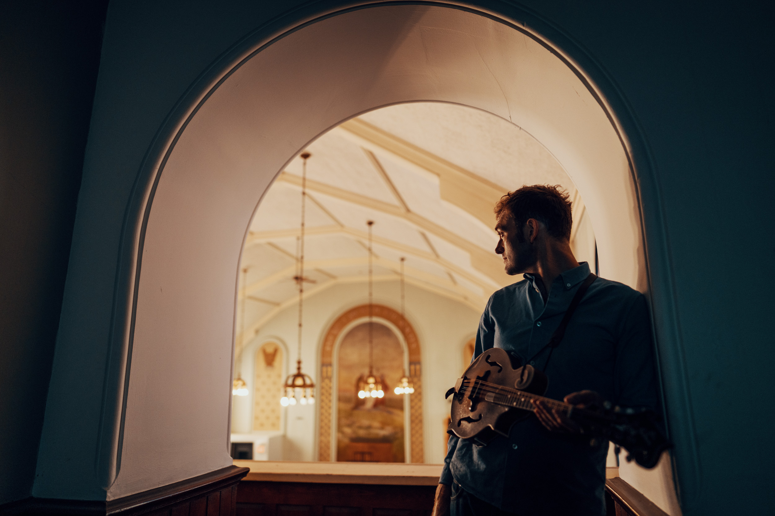Chris Thile standing in an archway holding his mandolin, looking away from the camera.