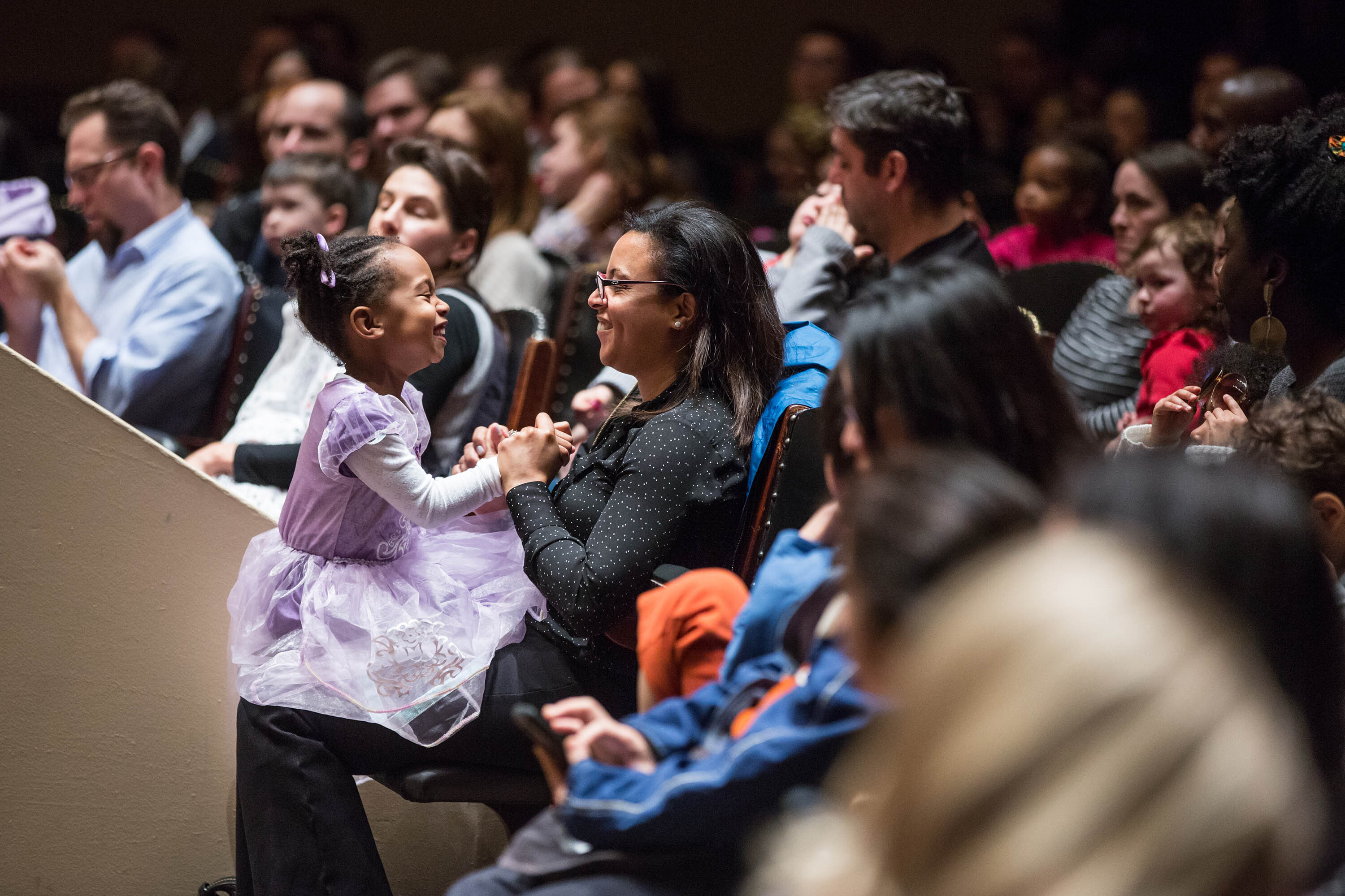 A young girl sits on her mother's lap and smiles at her in the audience.