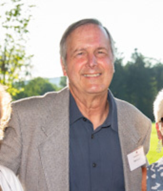 A headshot of a smiling man wearing a gray blazer and a blue button-down shirt, standing outside in front of trees