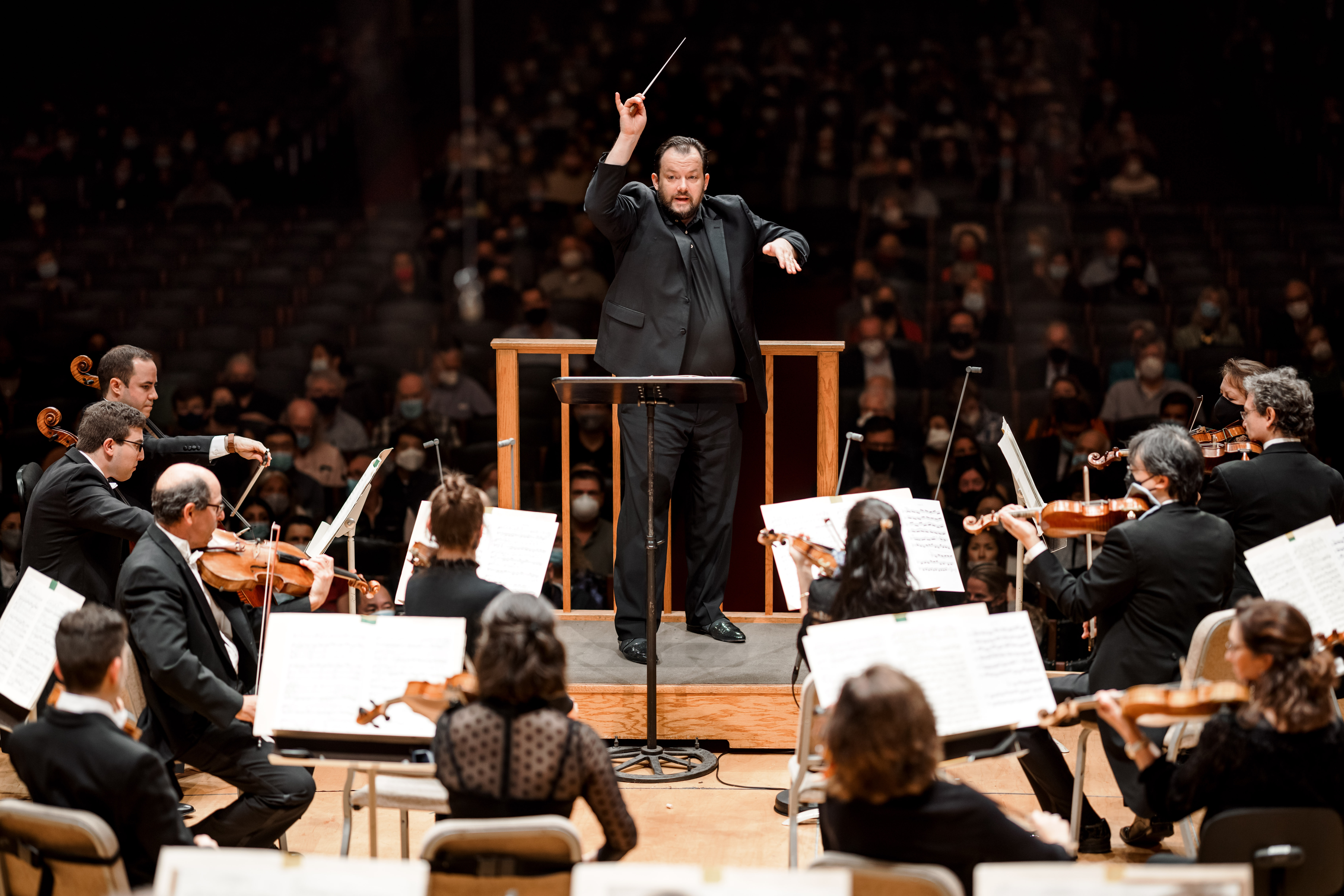 Man wearing black suit conducts orchestra onstage with audience behind him in darkness