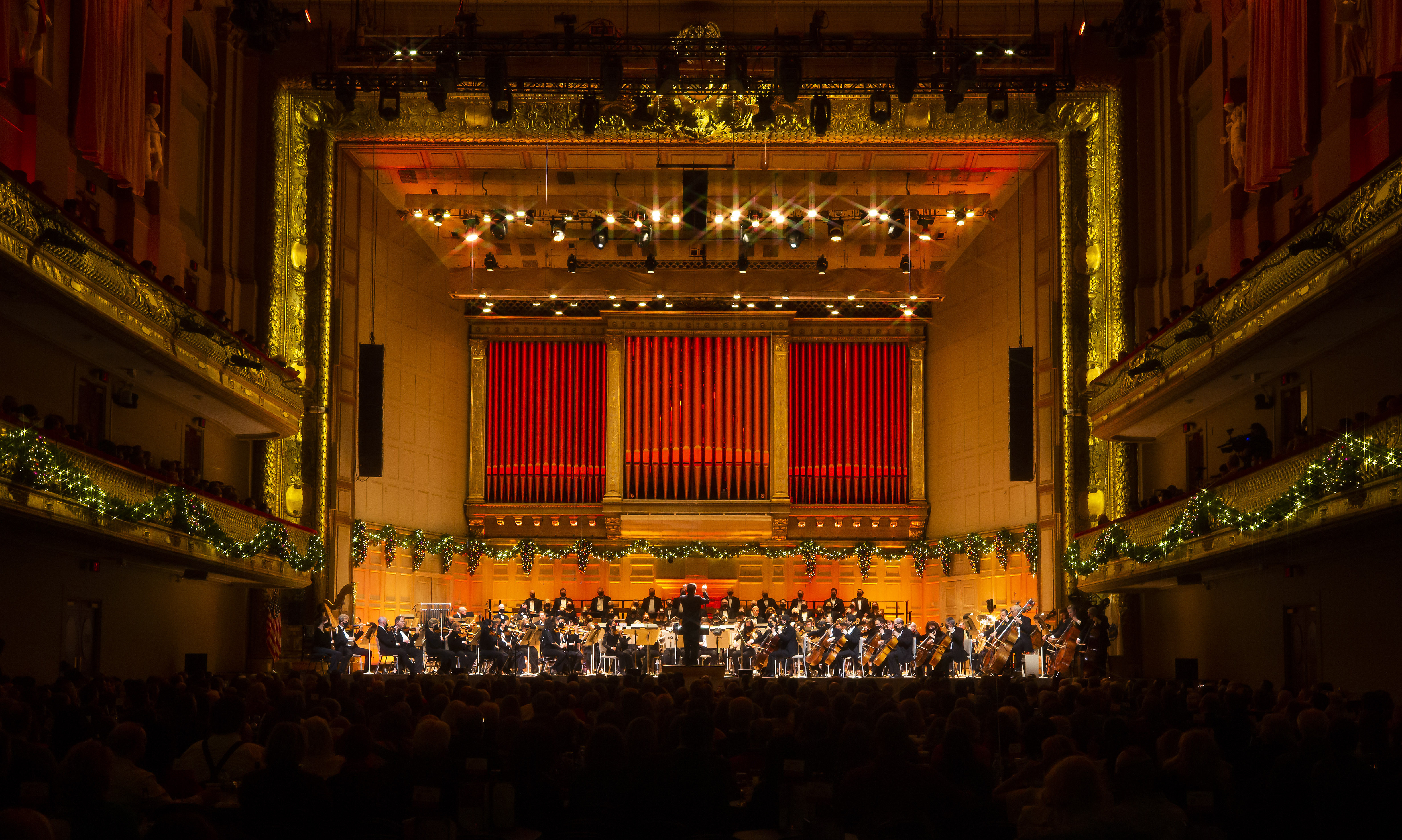 A full view of the stage of Symphony Hall, where Keith Lockhart stands in front of the orchestra as the stage is filled with gold and red lighting