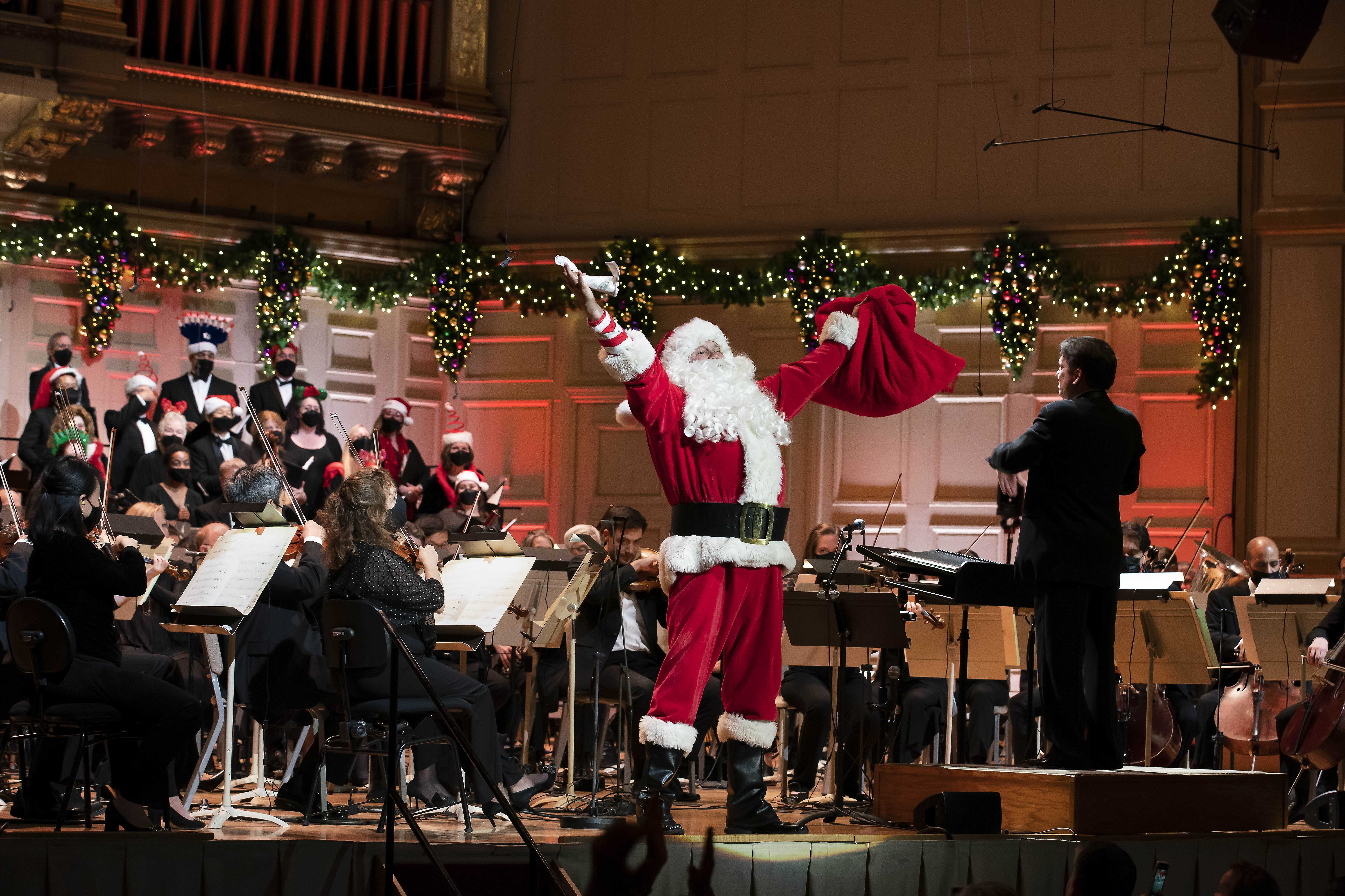 Santa Claus, with his arms outstretched, stands onstage of a decorated Symphony Hall, in front of the Boston Pops while Keith Lockhart conducts