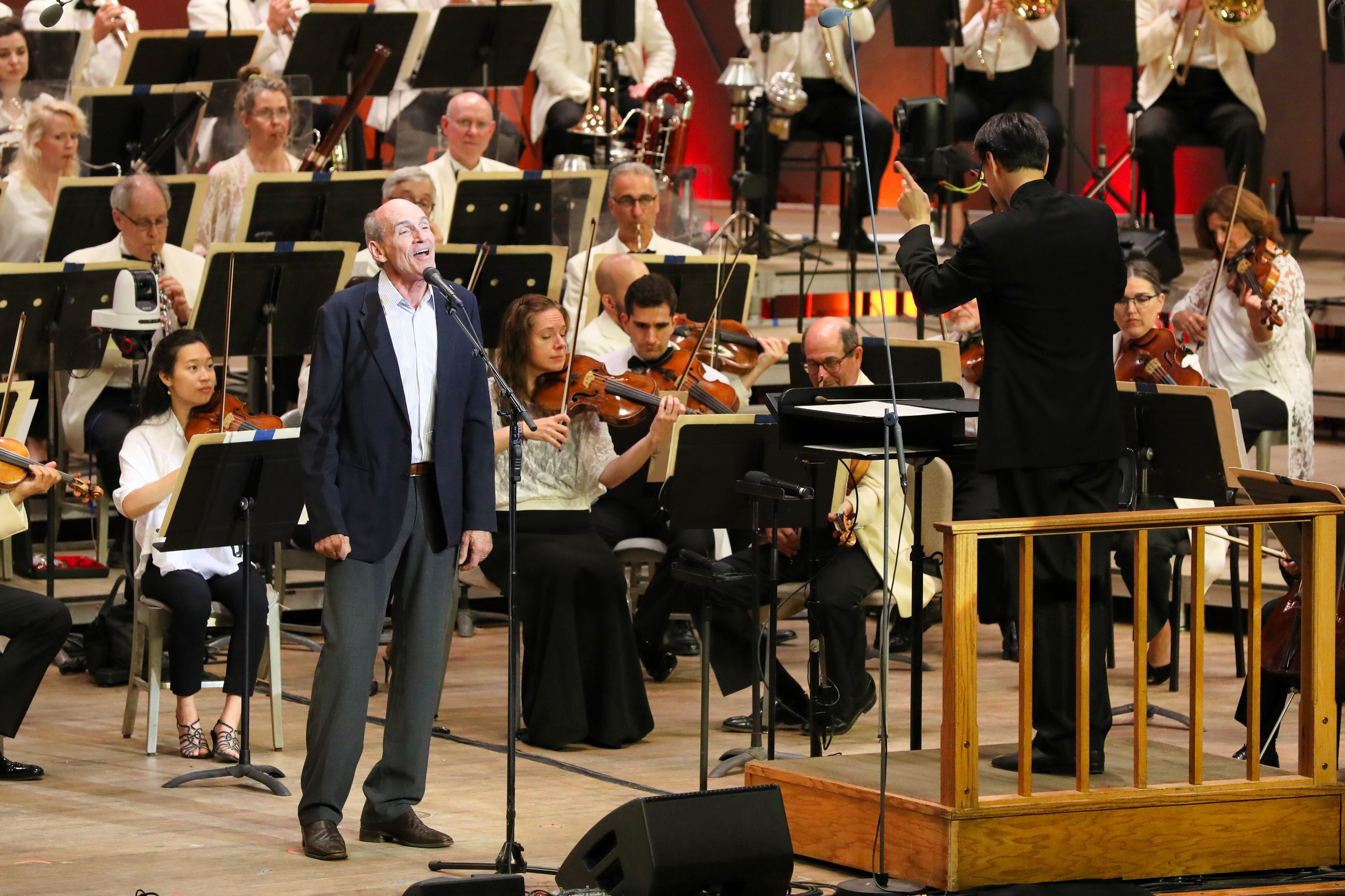 Balding man wearing navy blue suitcoat and gray pants stands at microphone while singing in front of orchestra onstage