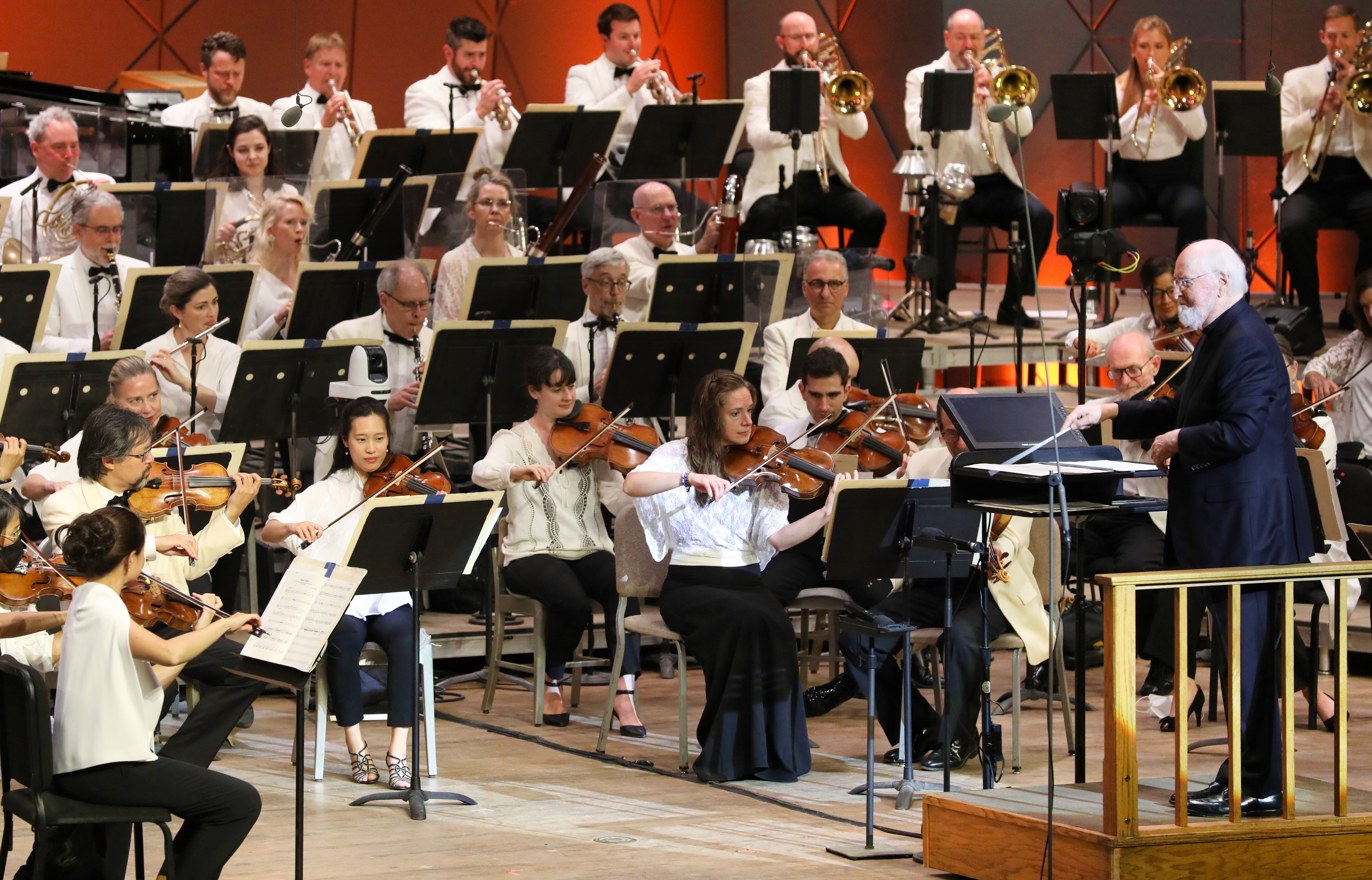 Older man stands at conductor podium in front of orchestra in the middle of playing onstage