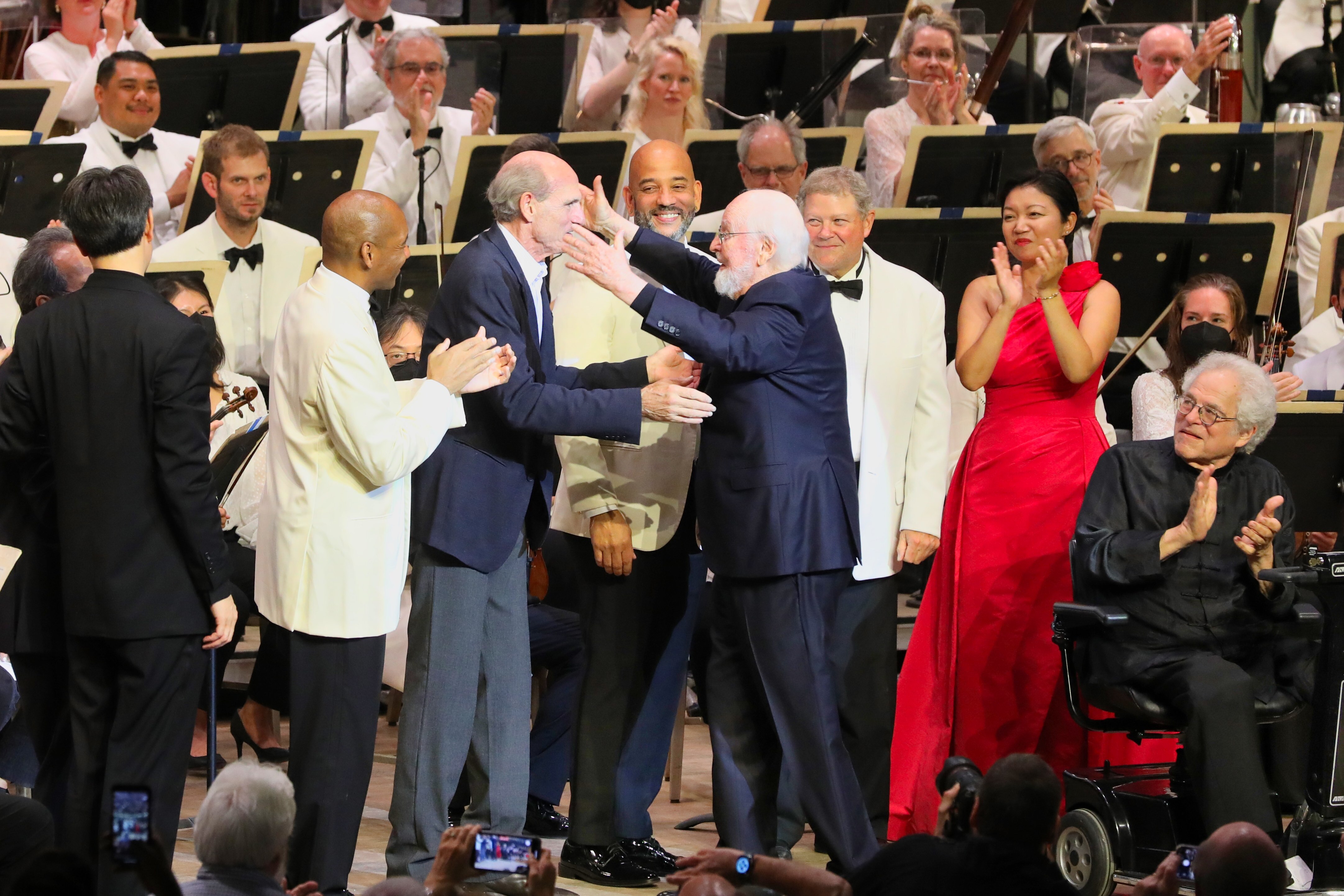 Older man wearing black suit opens arms to man while others smile and applaud as they look on