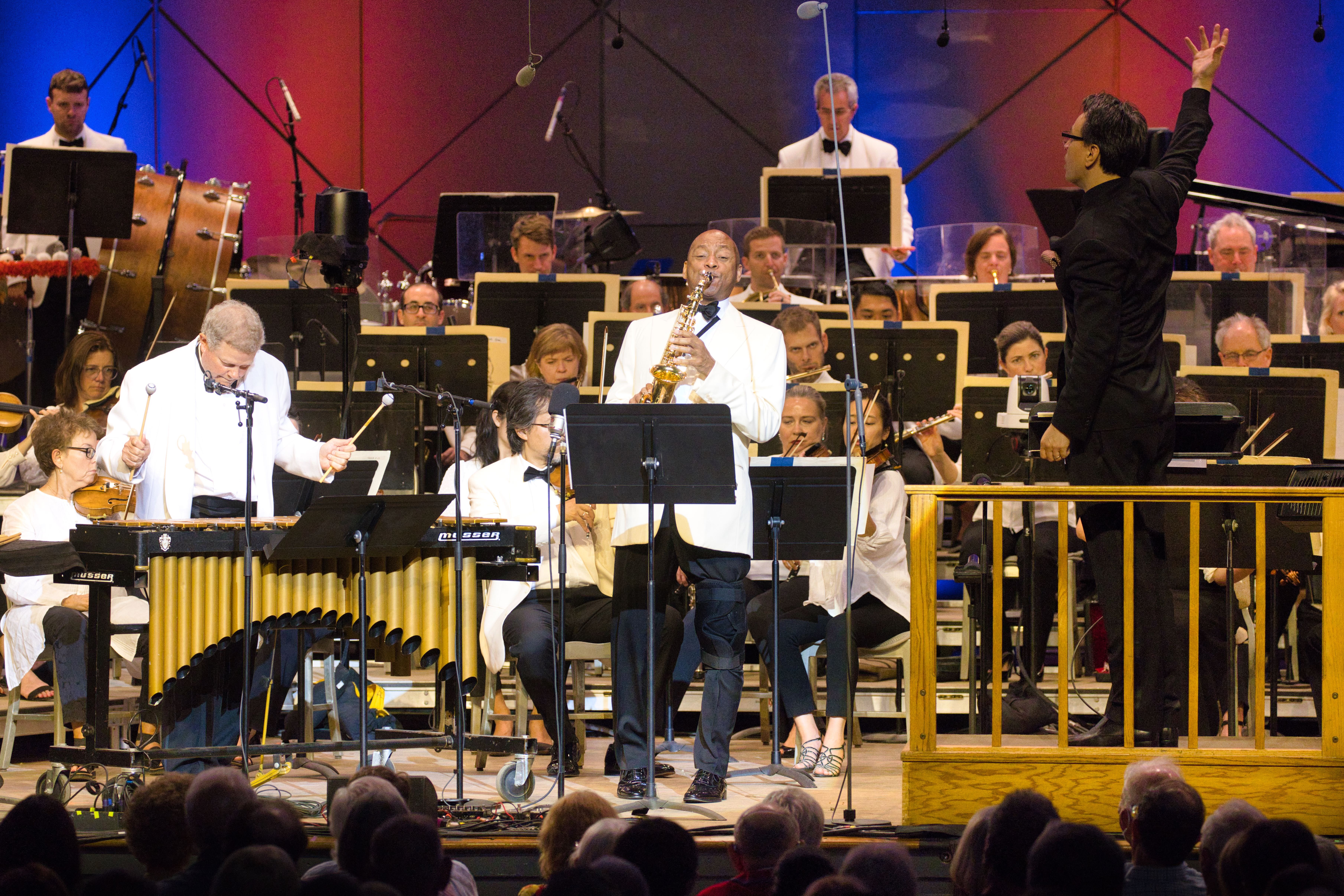 Conductor wearing black suit leads orchestra beside man wearing white suitcoat playing the saxophone and man wearing white suitcoat playing vibraphone