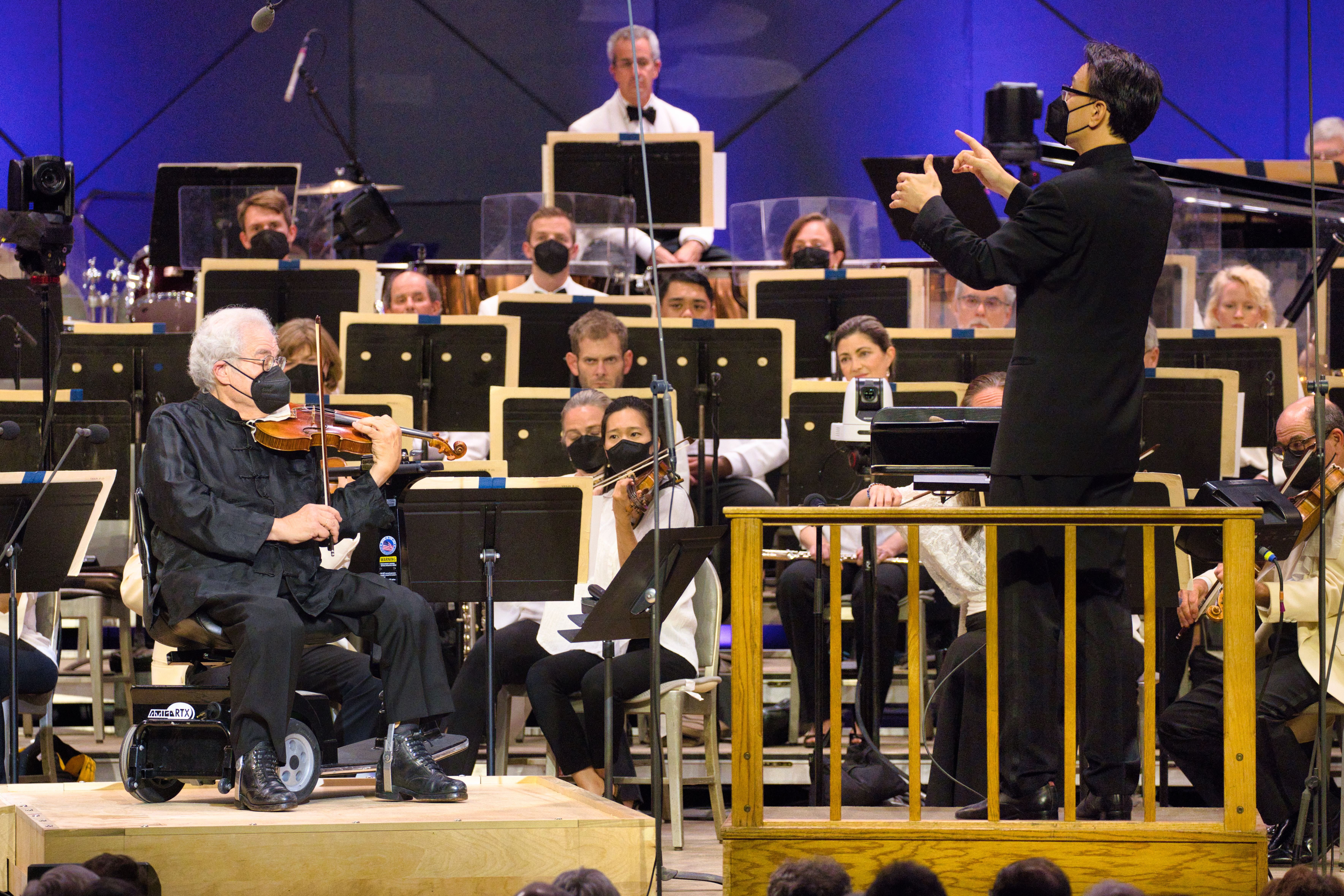 Man wearing black suit leads an orchestra onstage beside an older man sitting and playing the violin