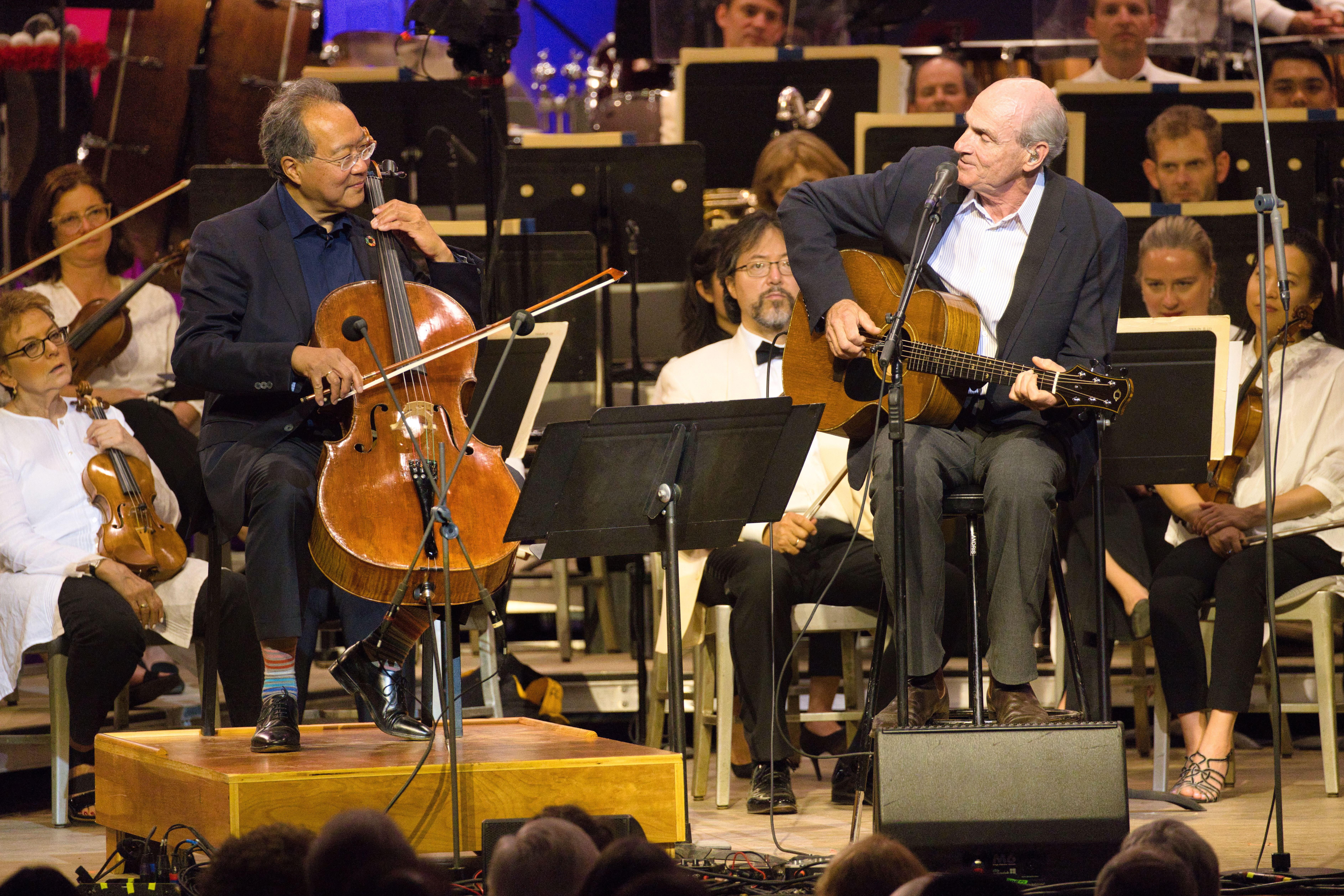 Man playing the cello performs alongside man playing the guitar, in front of an orchestra onstage