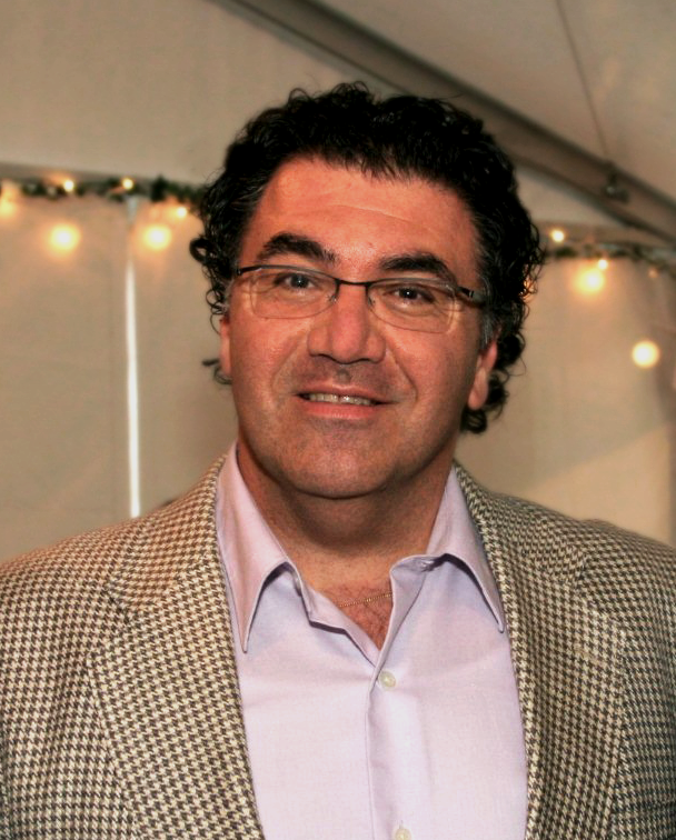 Man with curly black hair and glasses smiles, wearing a white button-down shirt and a tan jacket