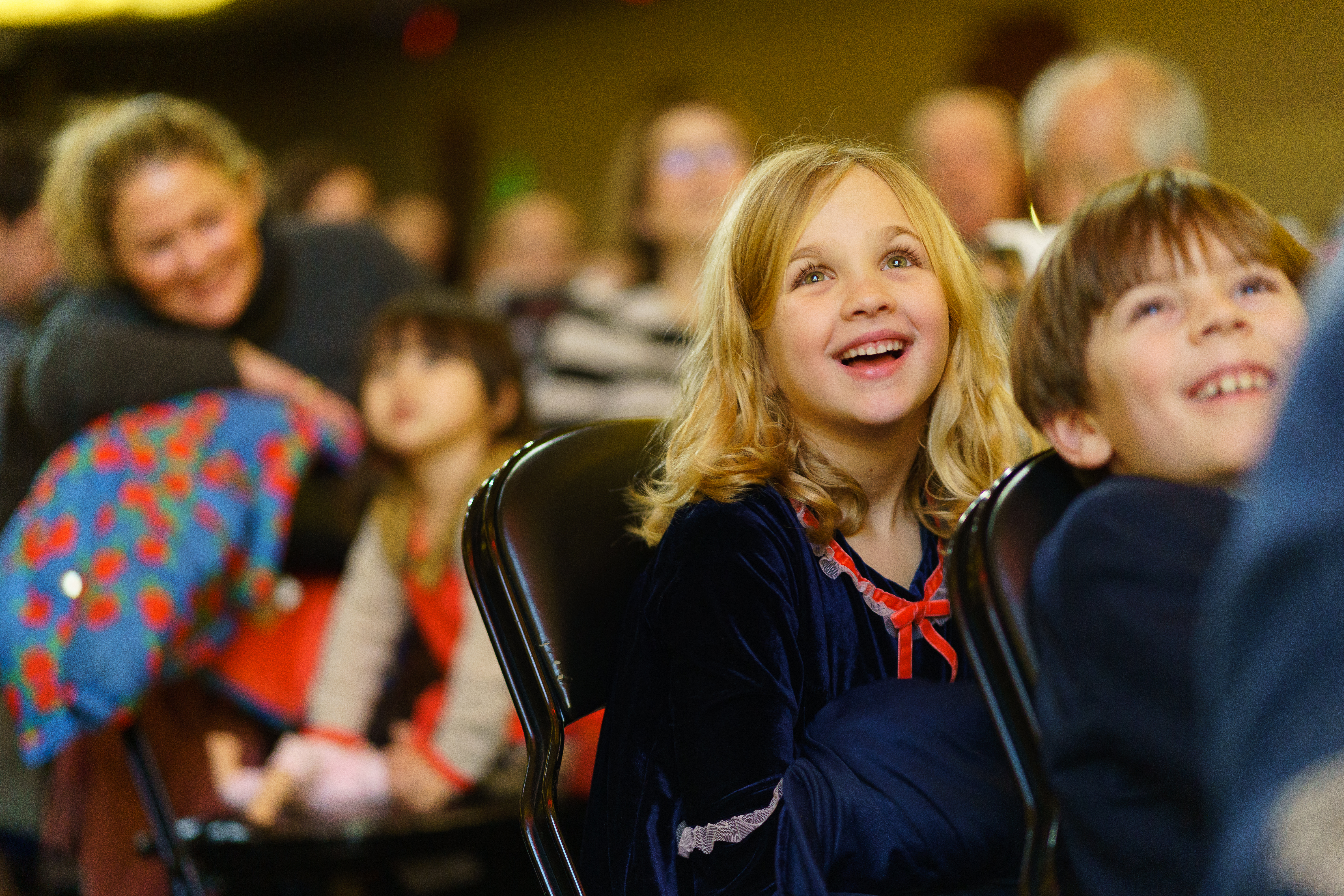 A young blonde girl and a young blonde boy stare ahead of them in awe, other audience members in Symphony Hall behind them watching in the same direction