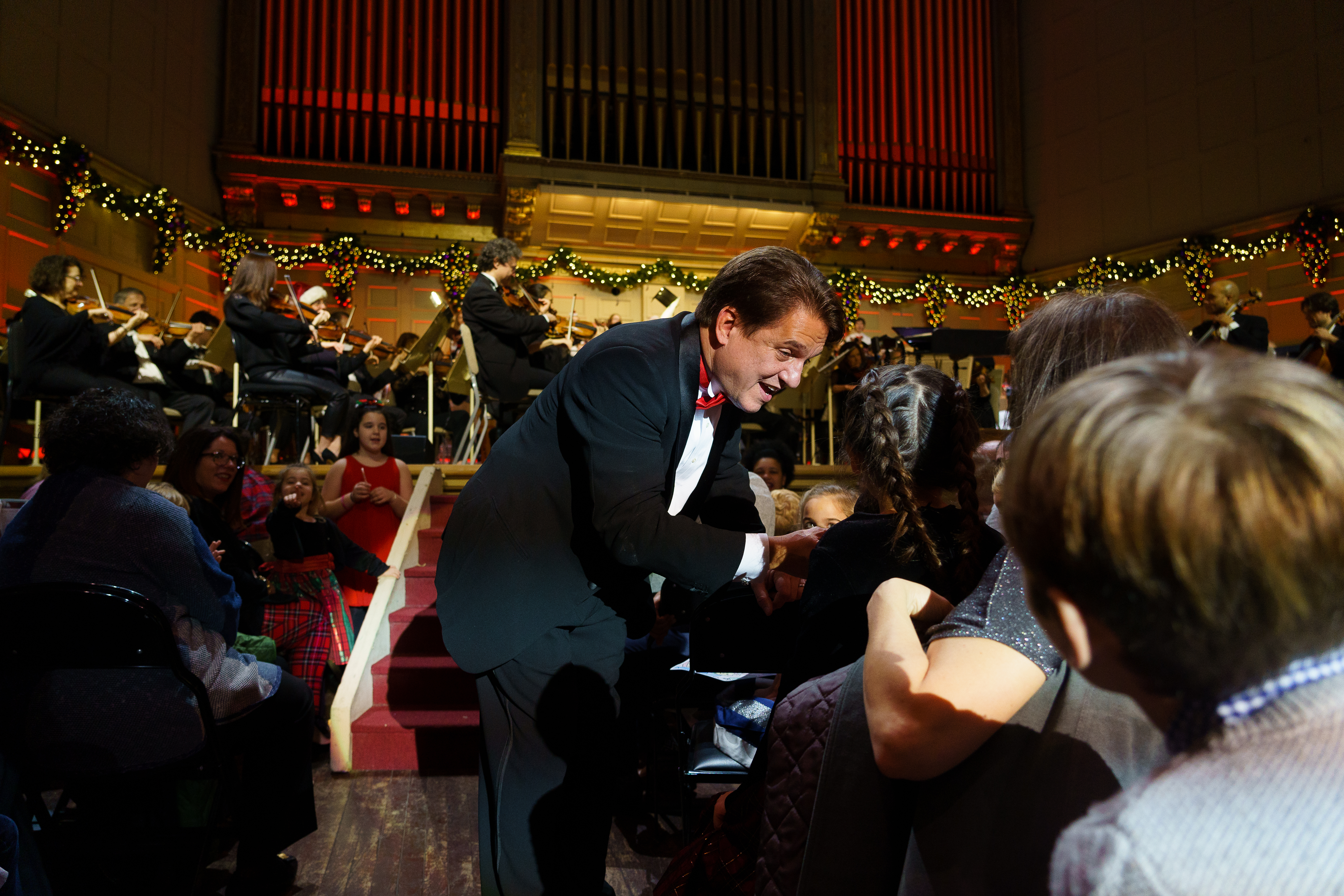 Keith Lockhart, wearing a tuxedo, leans over a young child in the audience, encouraging a singalong while the Boston Pops sits onstage behind him and performs.