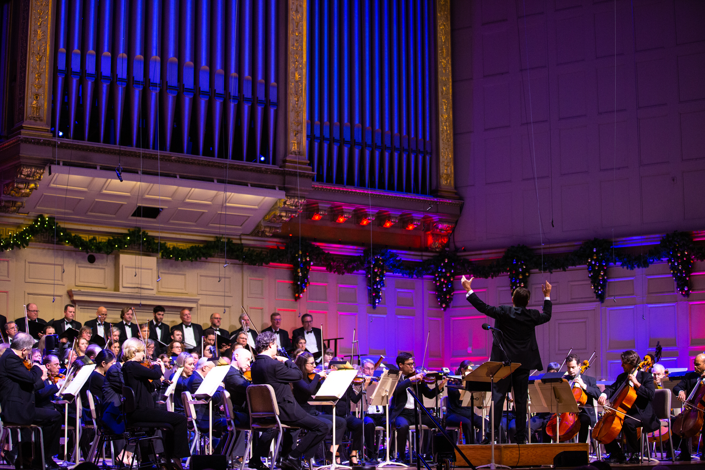 A view of the stage of Symphony Hall, adorned in lit garland and filled with blue and purple lights, as Keith Lockhart conducts the orchestra