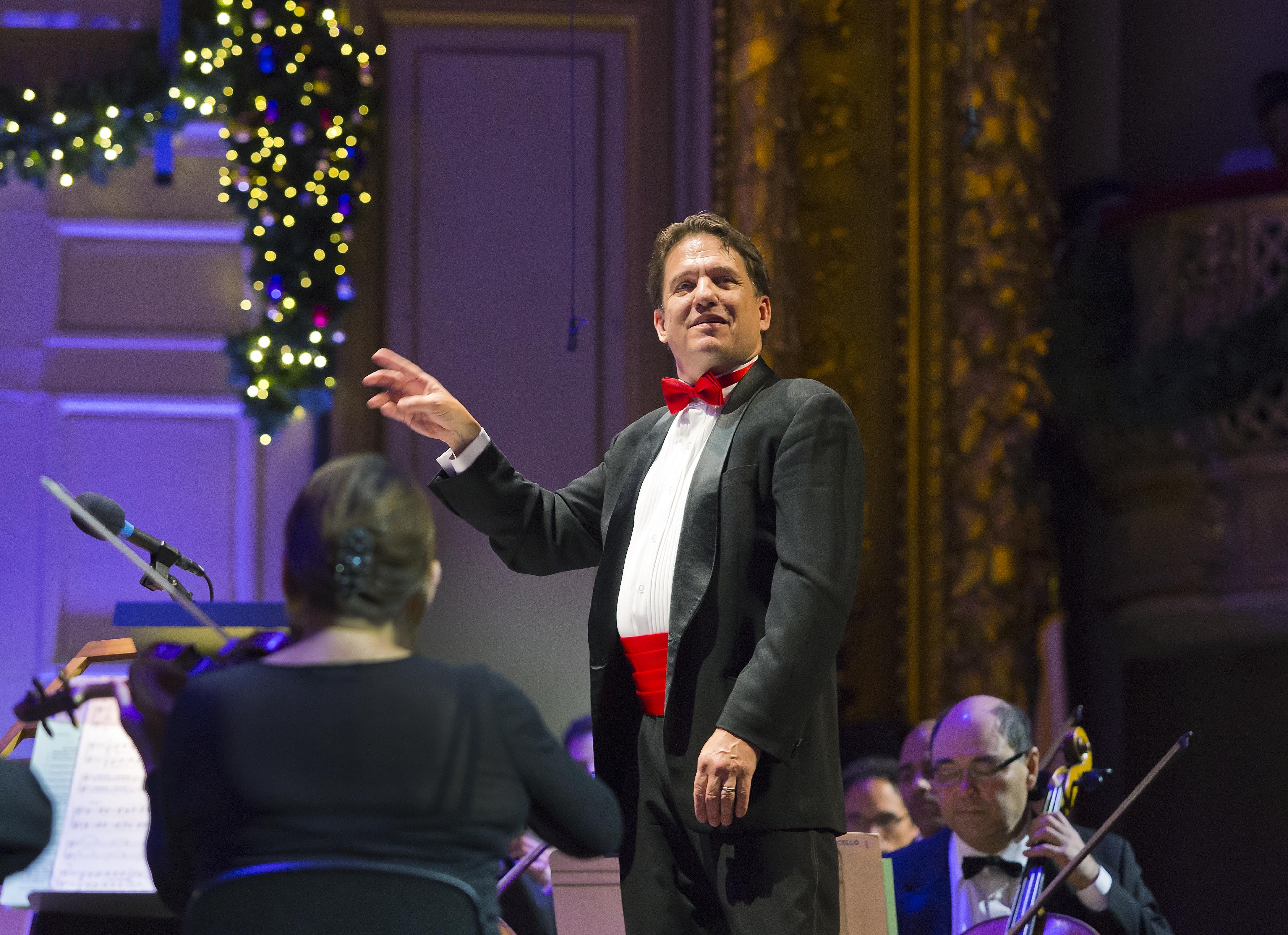 Keith Lockhart, wearing a tuxedo with a red belt and bow tie, stands on the decorated stage of Symphony Hall as he conducts the orchestra