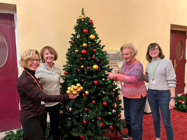 Four smiling volunteers stand beside a decorated Christmas tree in a hallway of Symphony Hall