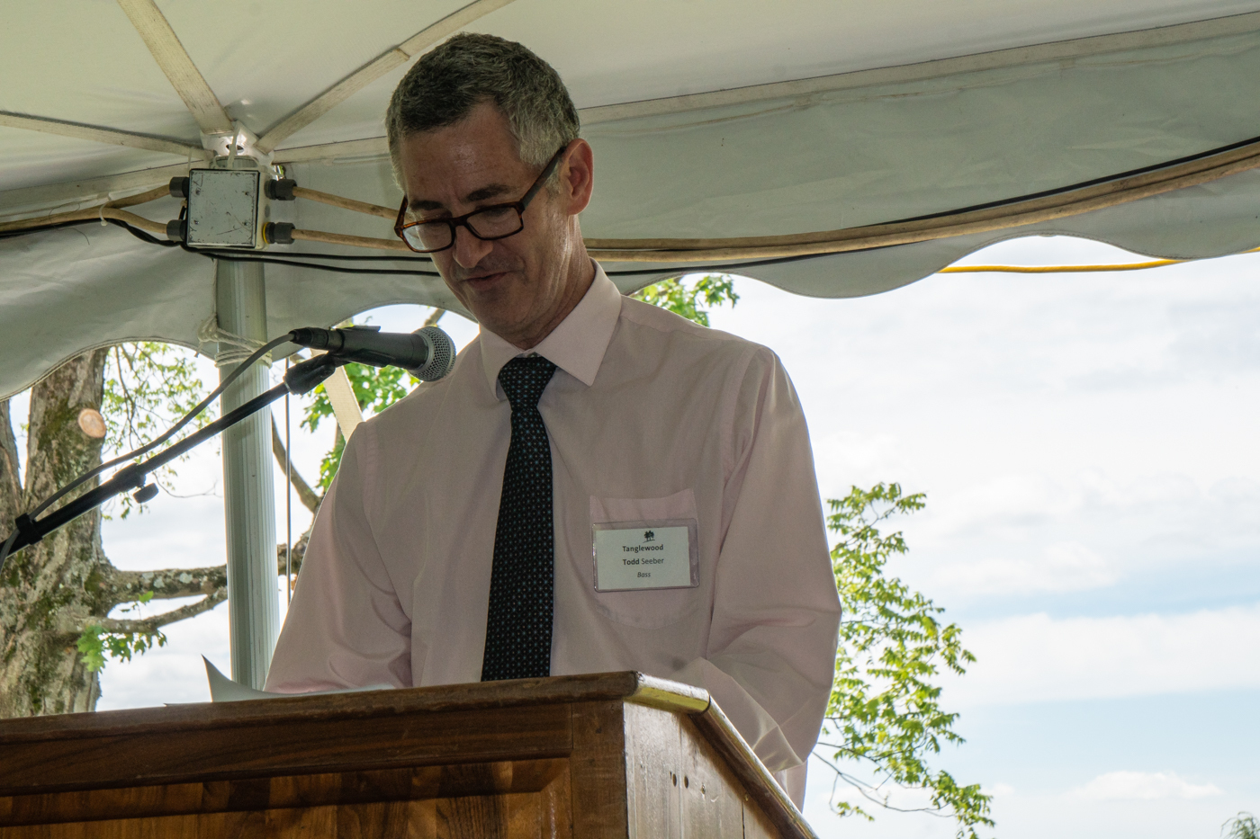 Man wearing glasses and a white shirt with a blue tie stands at a podium and speaks in front of a blue sky with trees behind him