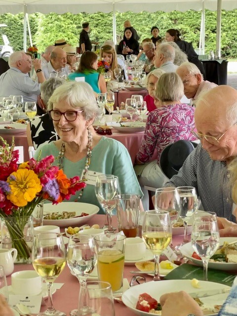 An older man and woman sit at a table covered in plates of food and glasses, in front of tables filled with other people eating, in front of green trees