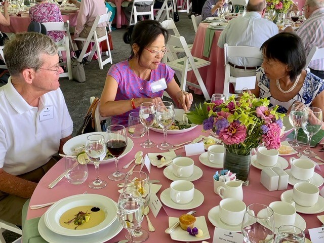 Two women and one man in the middle of eating at a table filled with plates chat with one another, with tables of others behind them