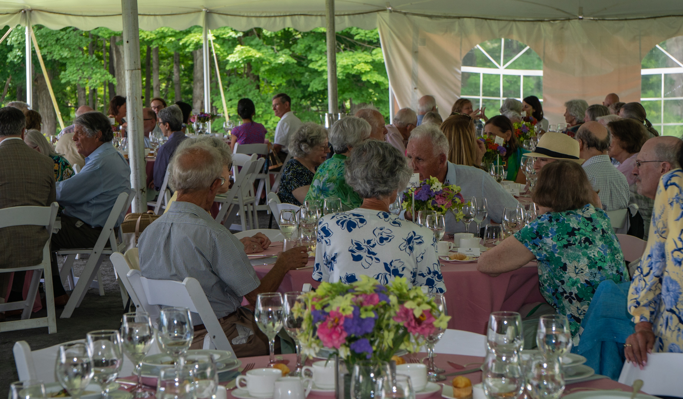 Tables filled with men and women are scattered around an indoor tent with windows and openings overlooking green trees