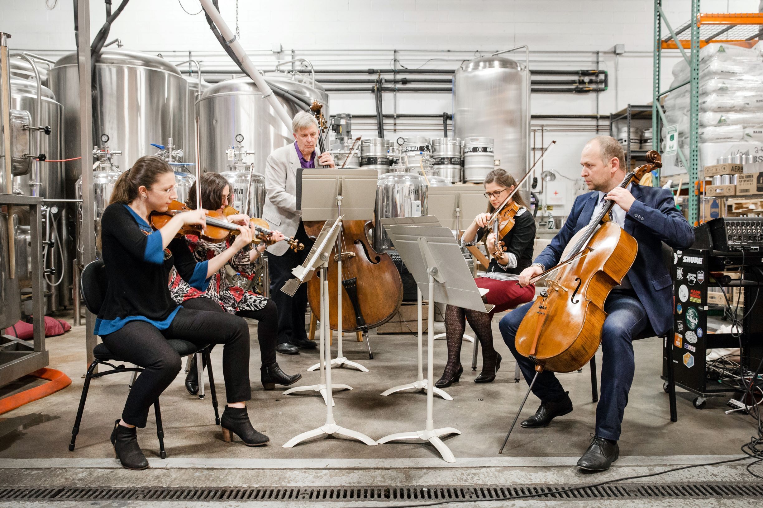A chamber group of string musicians perform at Aeronaut Brewery with industrial brewing equipment in the background.