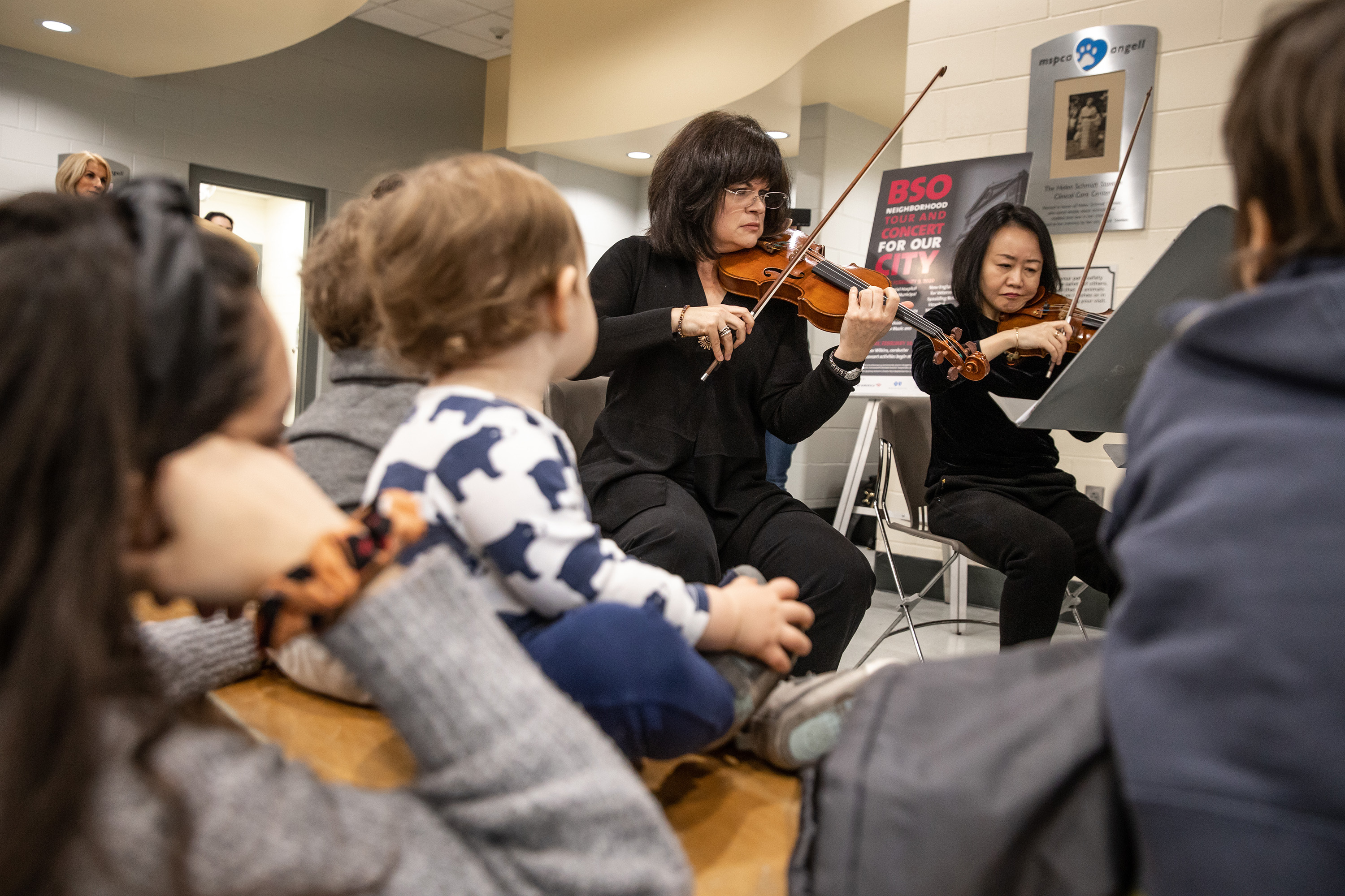 Catherine French has black hair and is dressed in all black as she plays violin, with a young child and a woman sitting and listening