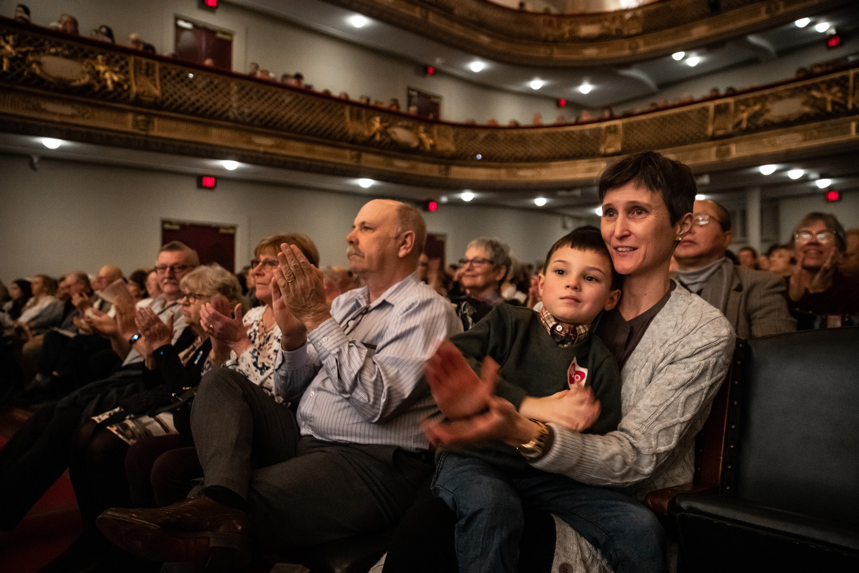A young boy sits on a woman's lap in Symphony Hall and the two clap