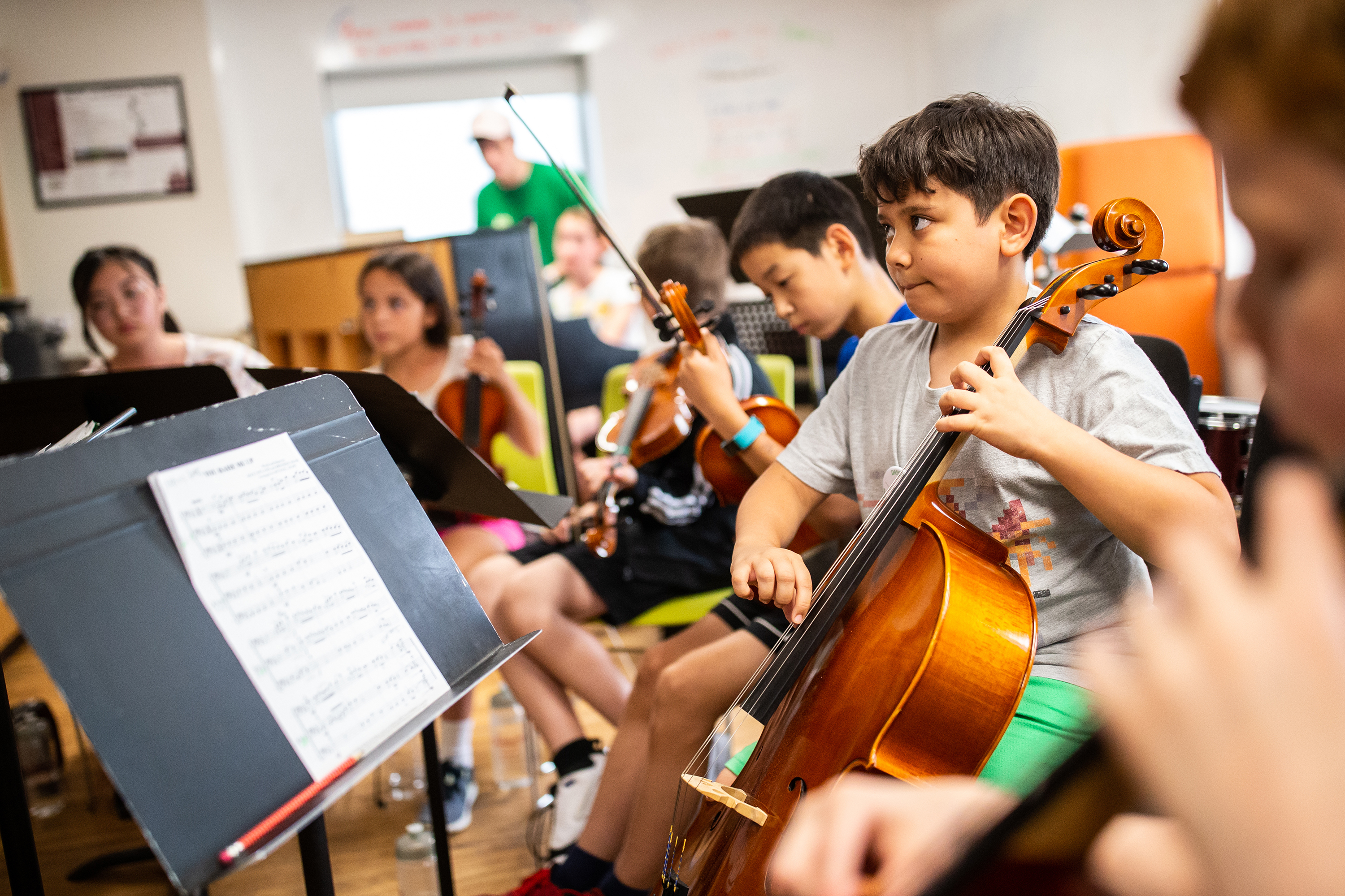A young boy in a grey t-shirt plays cello, with other young musicians sitting near him and holding their instruments