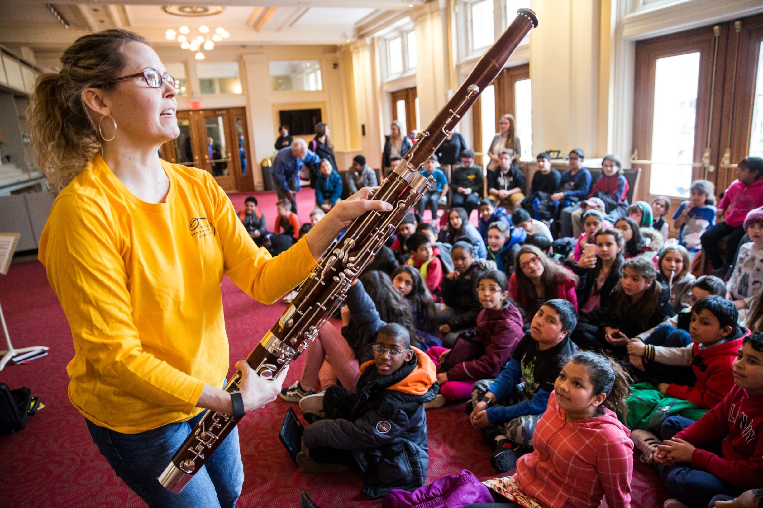 A musician wearing an yellow t-shirt holds up a bassoon. A group of school children watch and listen.