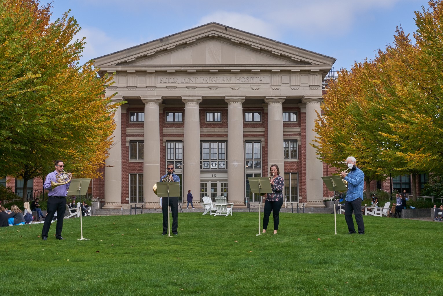A brass quartet stands spread out on a lawn playing music.