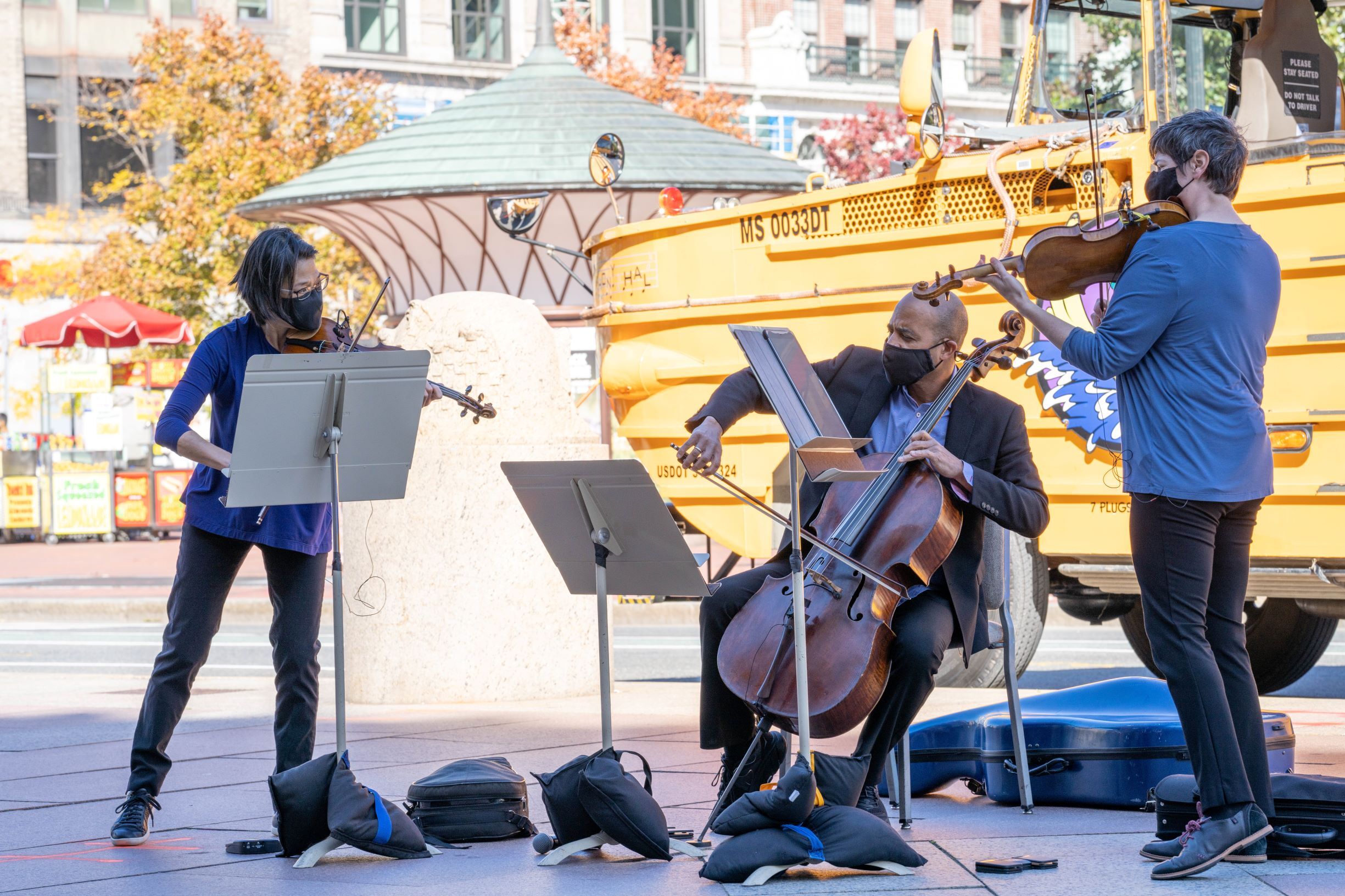 A string trio plays music in downtown Boston with a duck boat in the background.