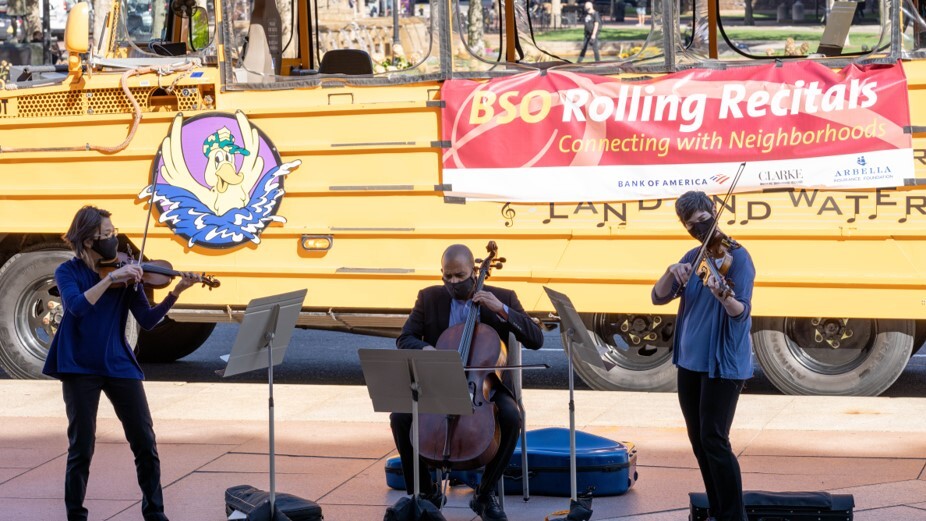 A city of Boston duck boat with a banner on it that reads "BSO Rolling Recitals" with musicians performing on the sidewalk in front of it.